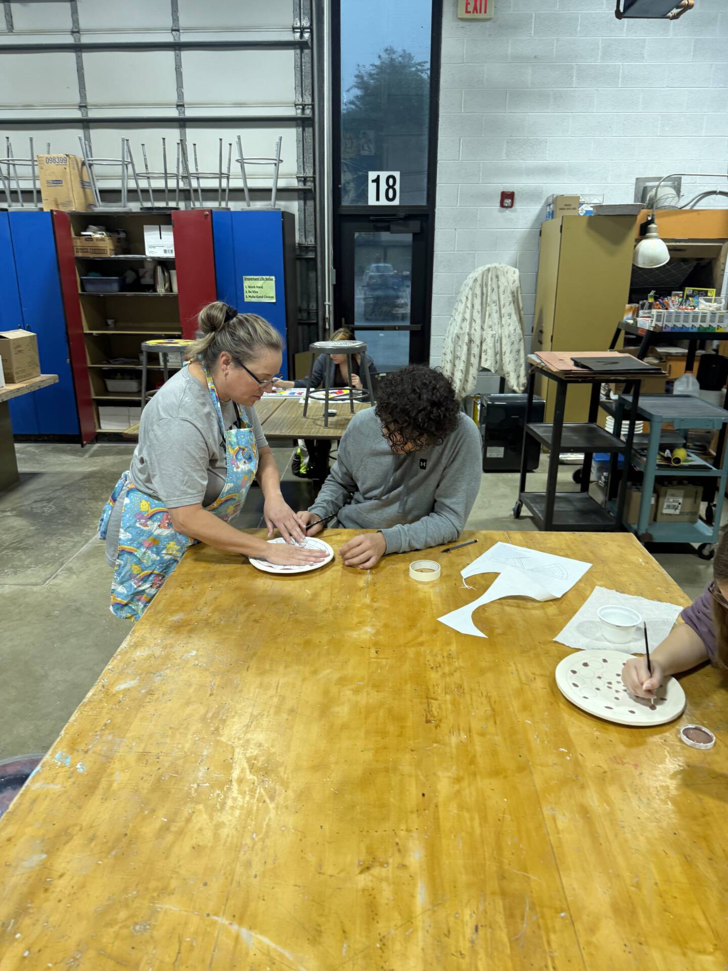 An adult assists a student with a hands-on art project at a workshop table while another student paints nearby.