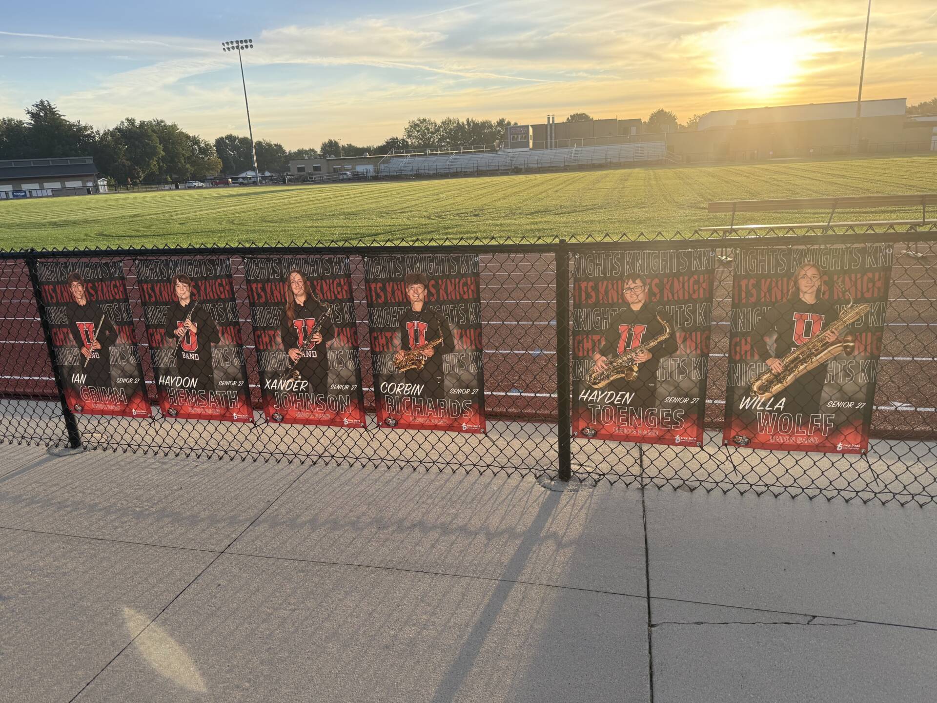 Senior band banners featuring six student musicians hang along a fence beside a sunlit athletic field.