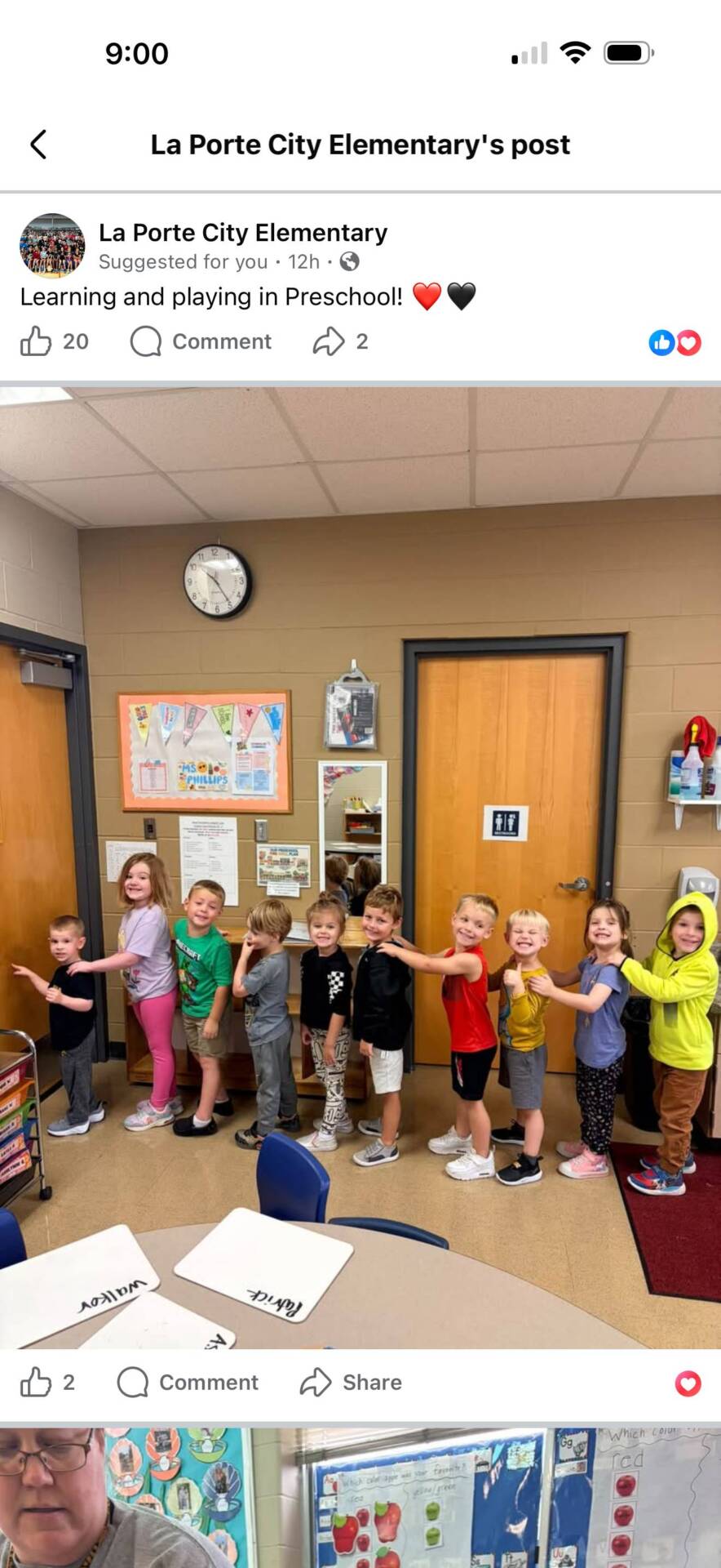 Nine preschool students line up behind one another in a classroom, smiling and holding onto each other's shoulders.