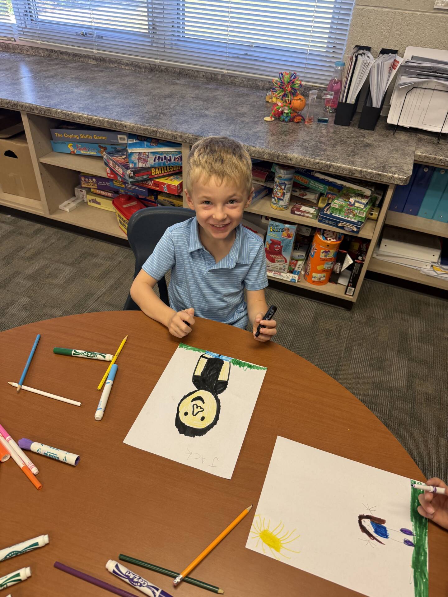 A smiling young boy in a blue striped shirt sits at a table displaying his colorful drawing and art supplies.