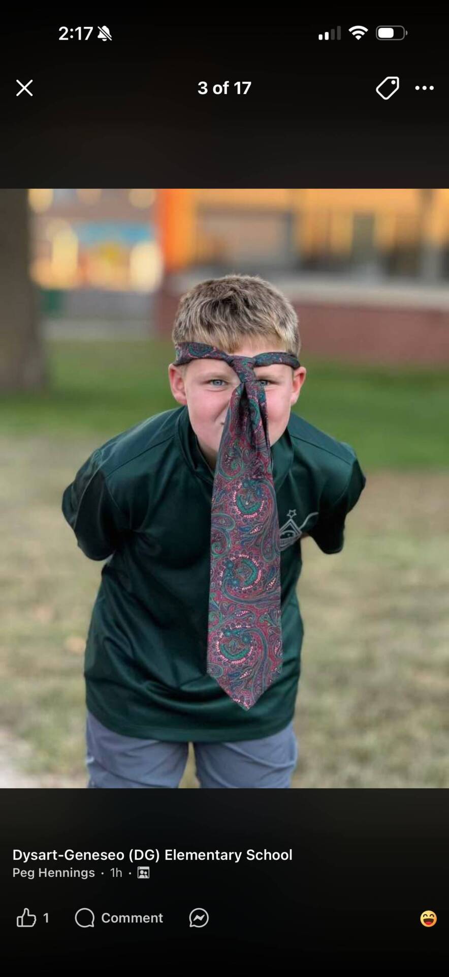 A smiling student outdoors wears a dark green shirt and a patterned necktie tied around his head.