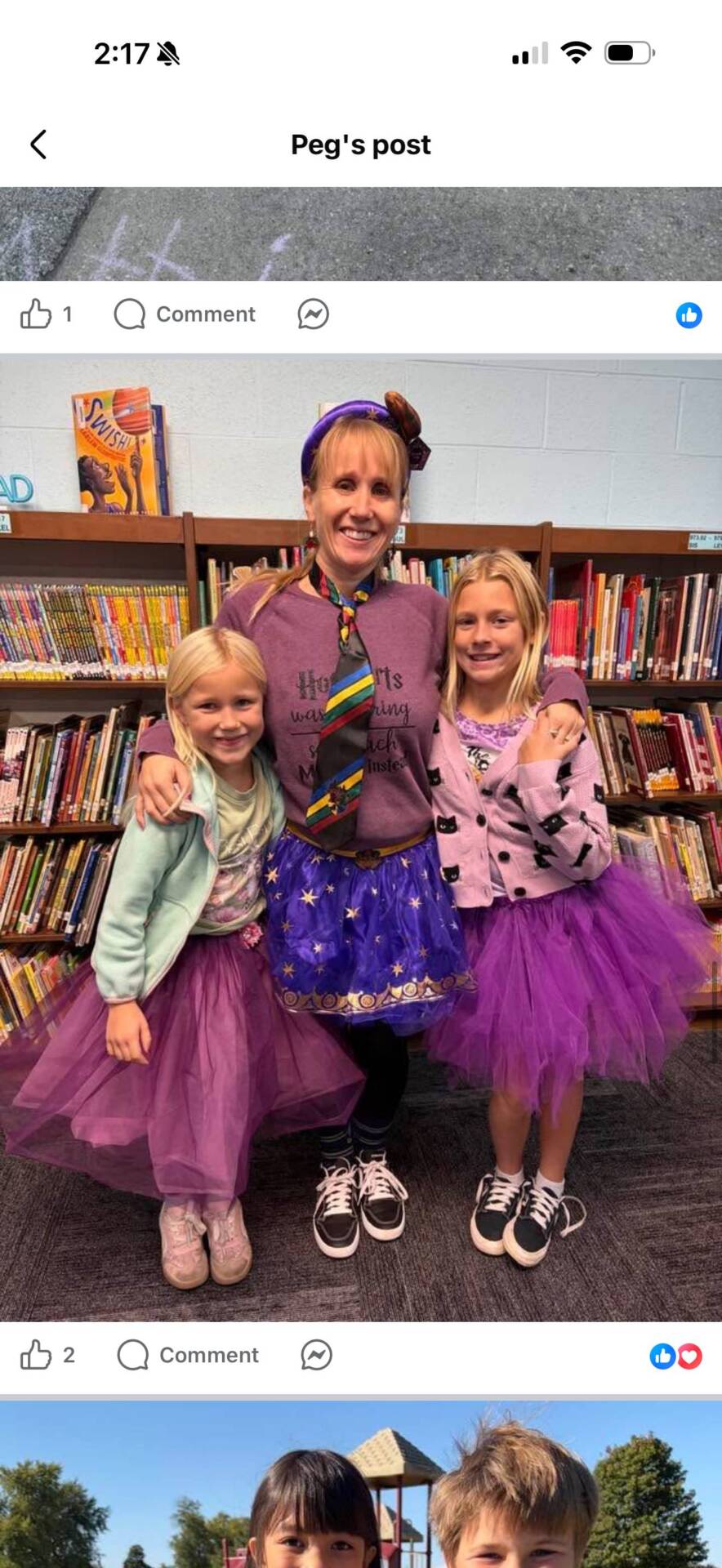 A teacher and two smiling students wearing colorful purple tutus pose together in front of library bookshelves.