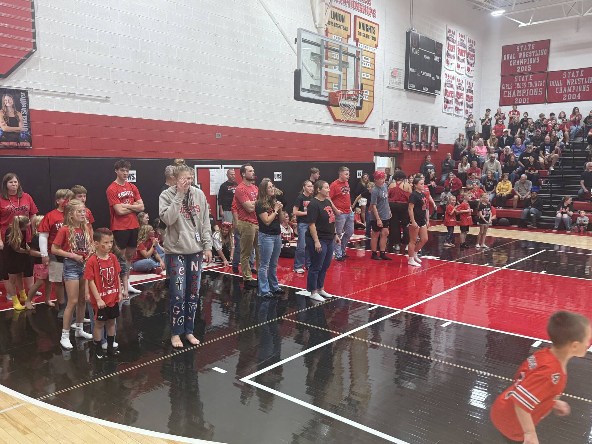Students, families, and staff gather on the gym floor and bleachers for a school spirit assembly, dressed in red and black.