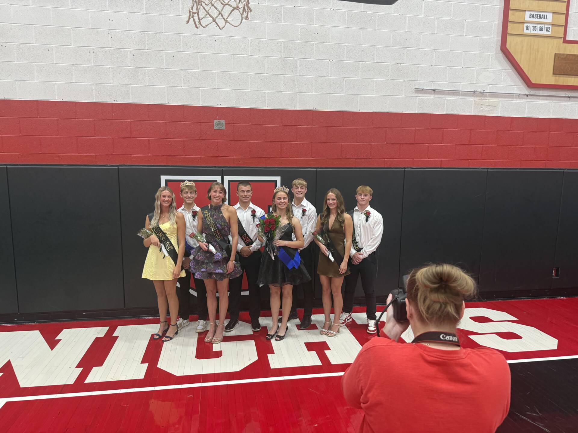 Eight homecoming court members wearing crowns, sashes, and formal attire pose for a photo in the school gym.