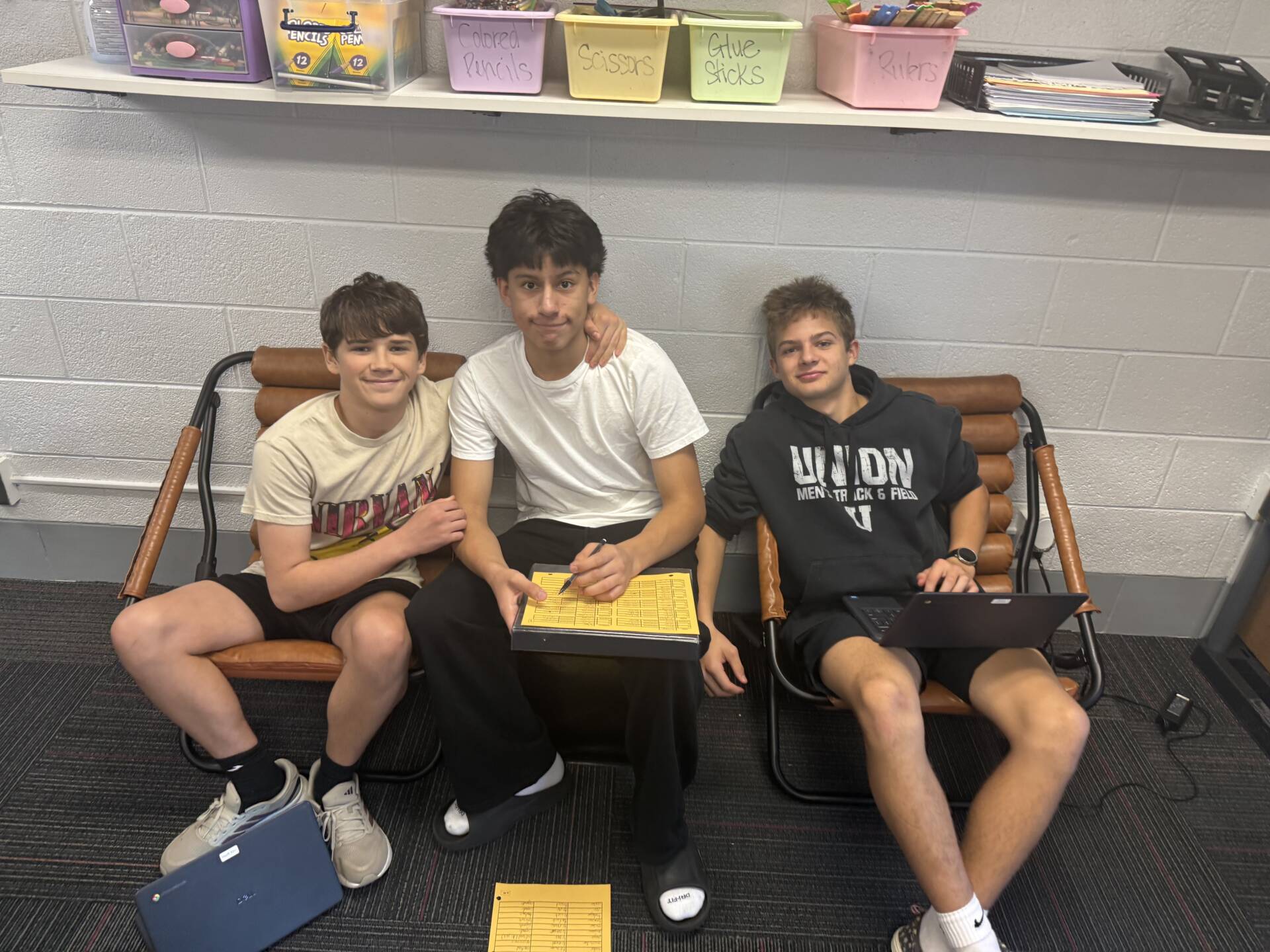 Three students sit together in a classroom with laptops and paperwork, smiling for the camera.