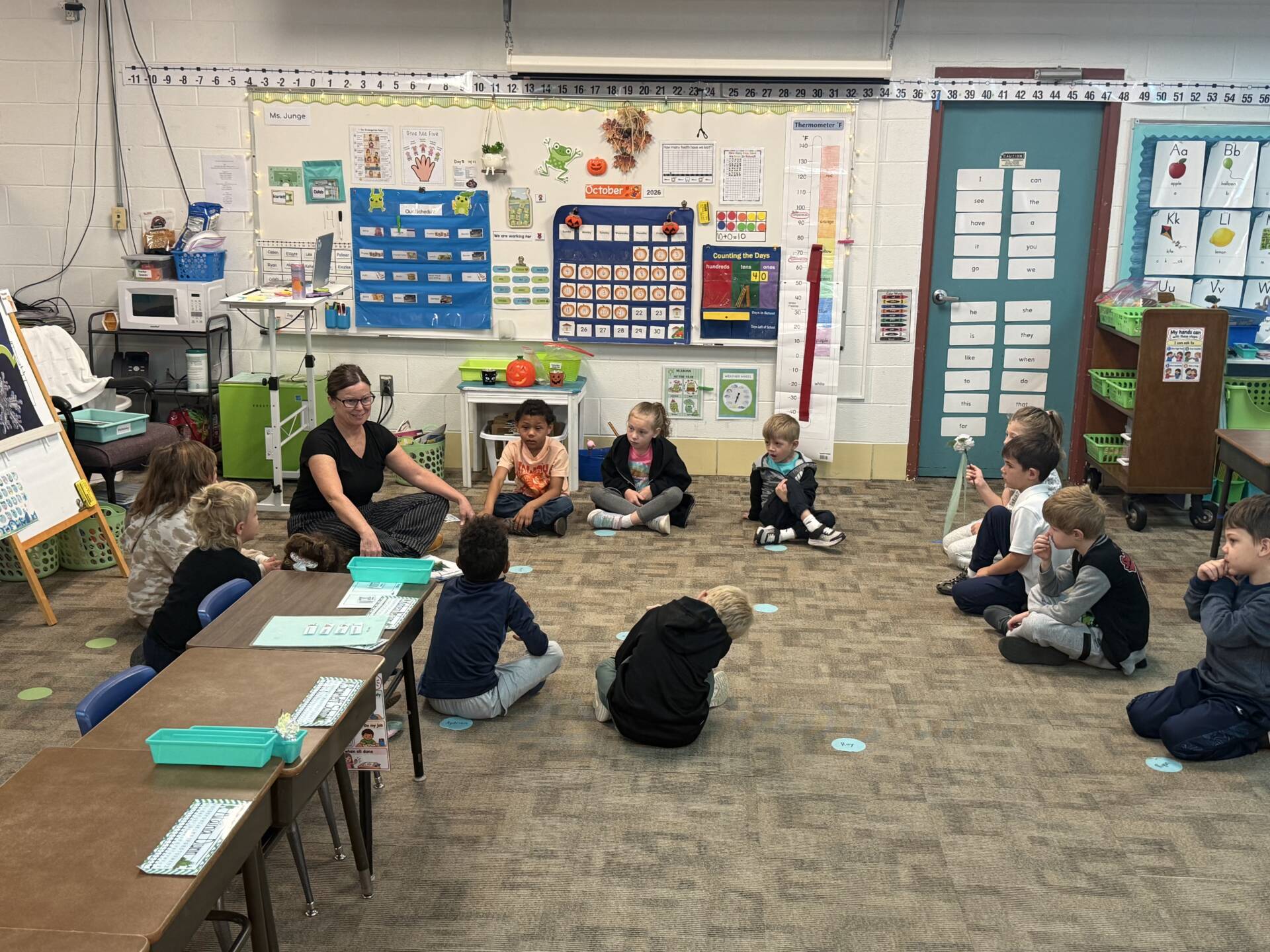 An elementary teacher sits on the carpet with students gathered in a circle for a group lesson in a colorful classroom.