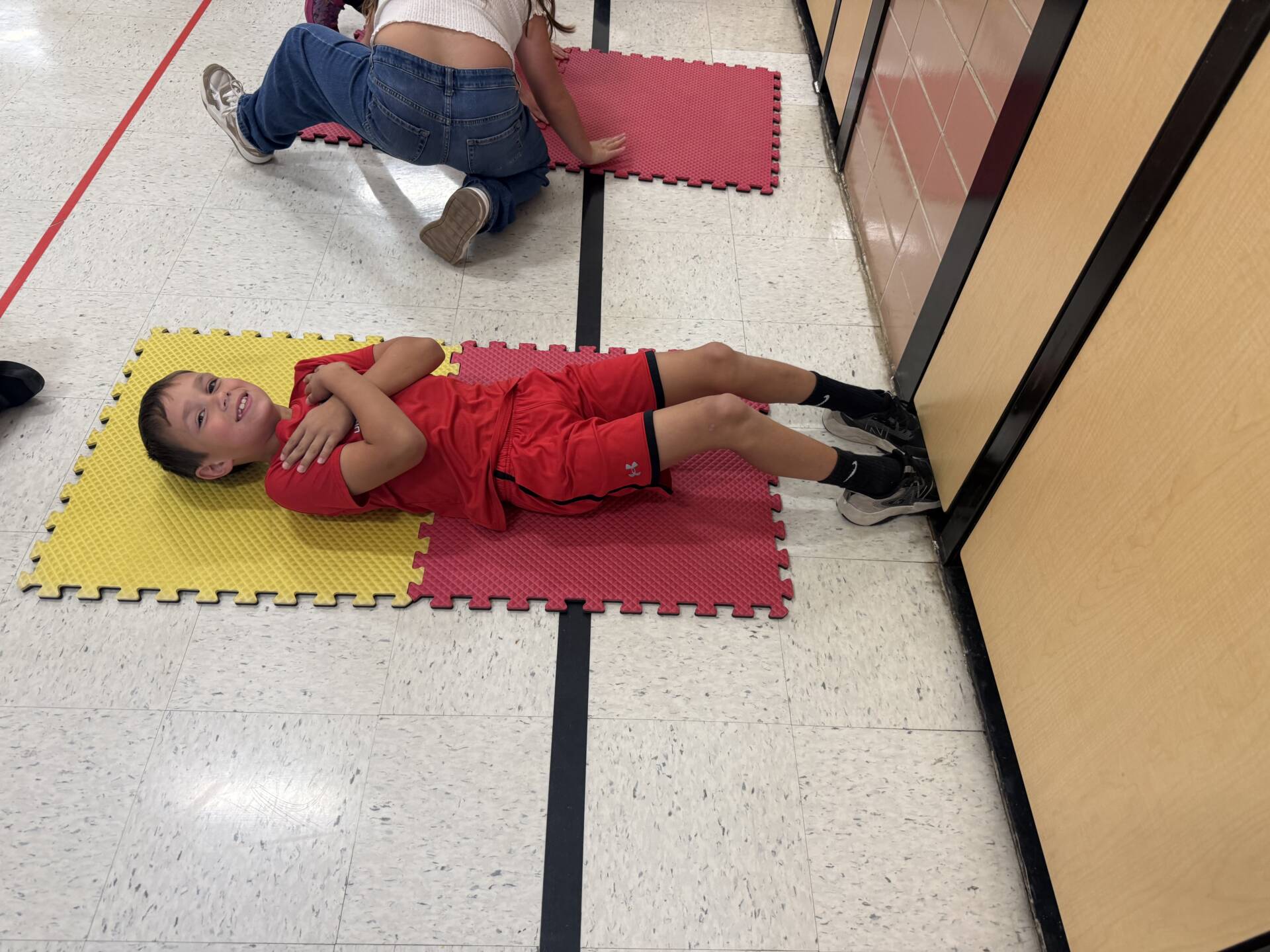 A smiling student in red athletic clothes lies on colorful foam mats during an indoor physical education activity.