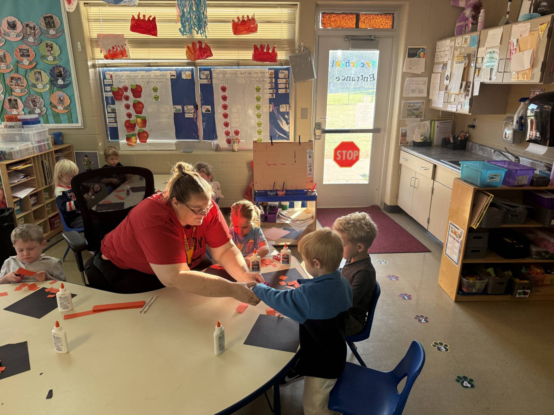 A teacher in a red shirt assists preschool students with orange and black paper crafts at a classroom table.