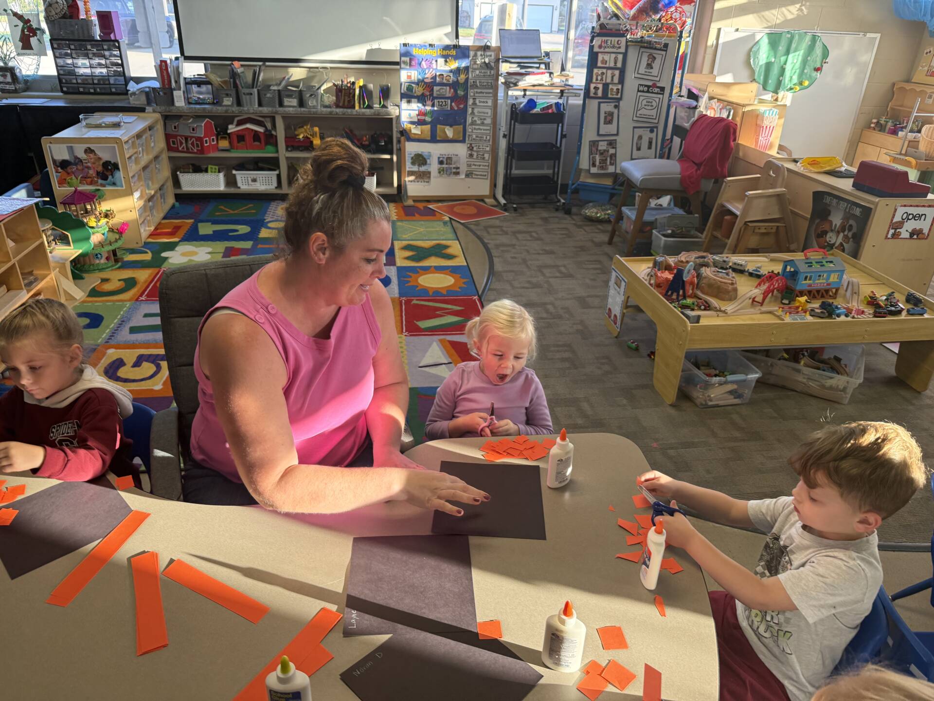 A teacher in a pink shirt helps preschoolers create paper crafts using orange strips, scissors, and glue in a colorful classroom.