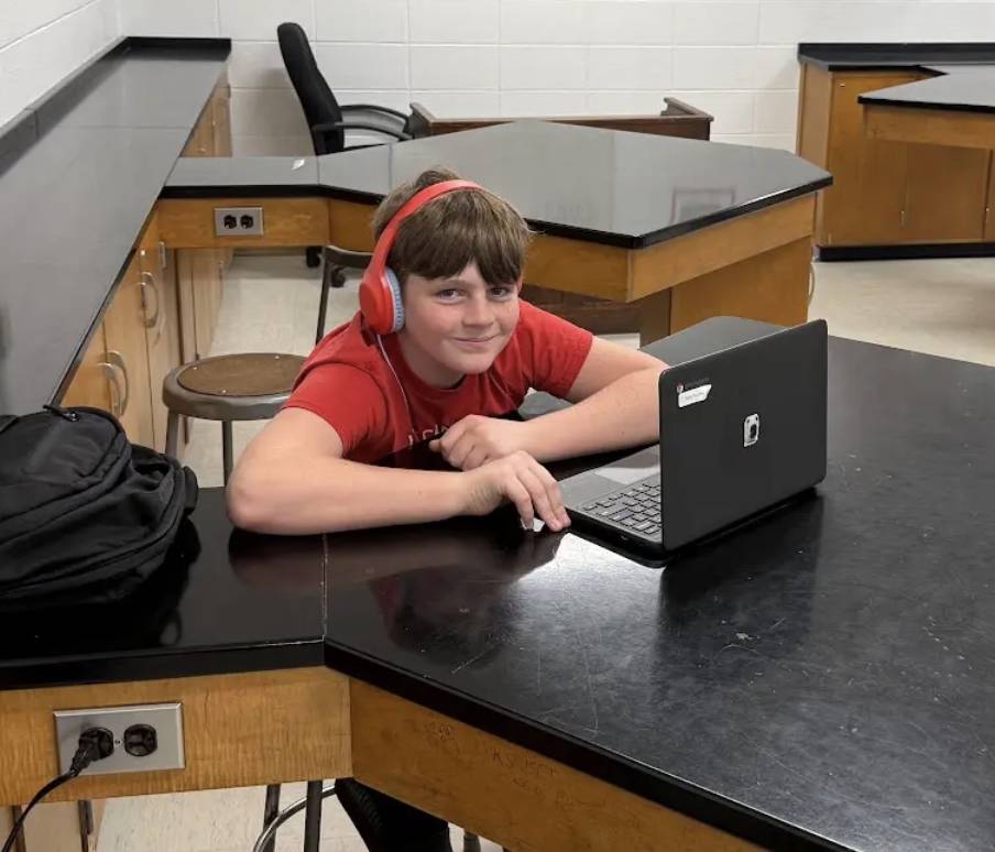 A smiling student wearing red headphones works on a laptop at a science classroom lab table.