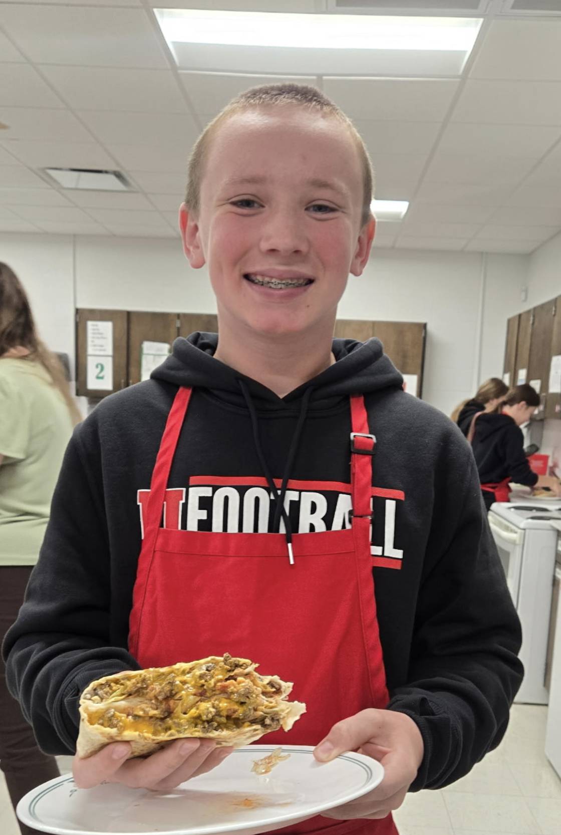 A smiling student in a red apron holds a plate with a freshly prepared stuffed tortilla in a classroom kitchen.