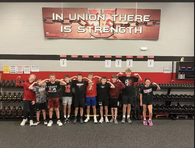 Ten students pose and flex their arms in a school weight room beneath a banner reading “In Union There Is Strength.”