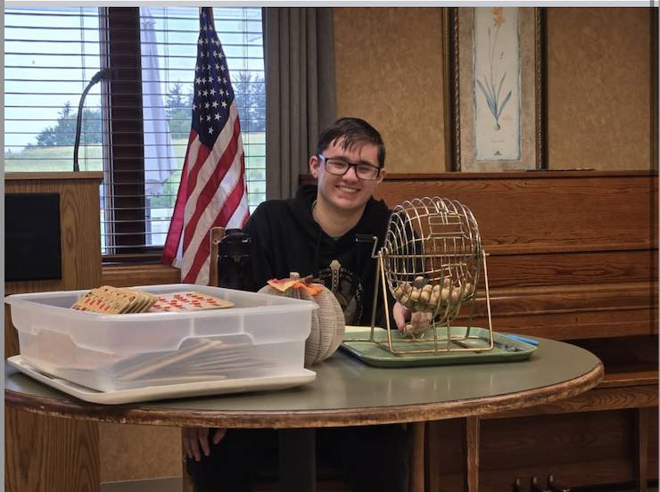 Smiling student sits behind a table with a bingo cage, cards, and supplies, with an American flag in the background.