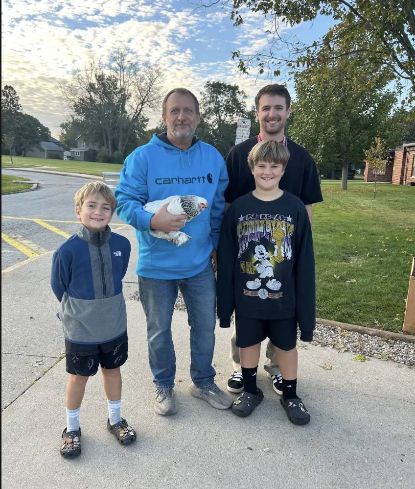 Four smiling people pose outdoors, including a man in a blue sweatshirt holding a white chicken.