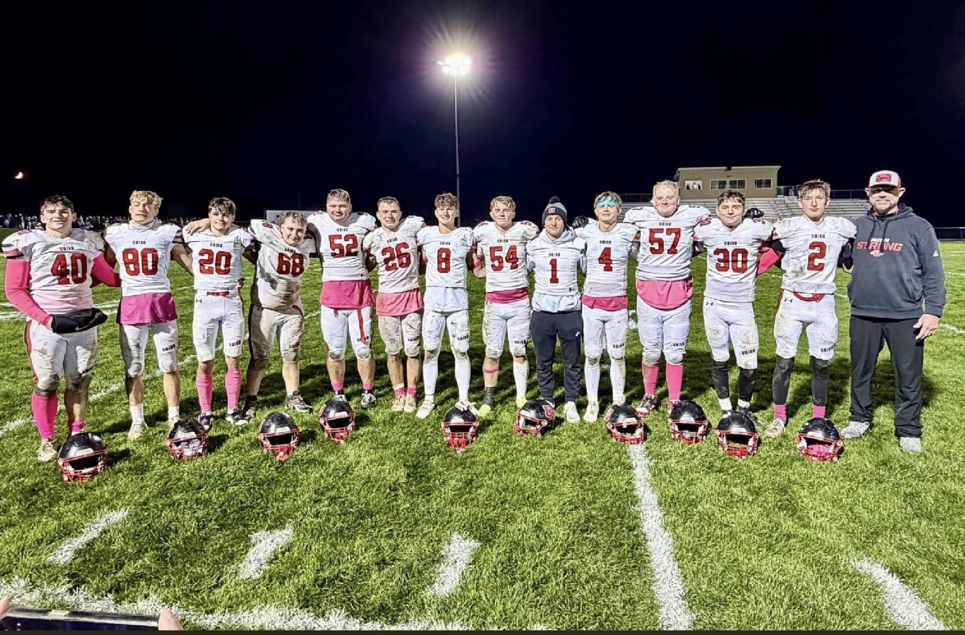 High school football players in white uniforms with pink accents pose alongside their coach under stadium lights after a nighttime game.