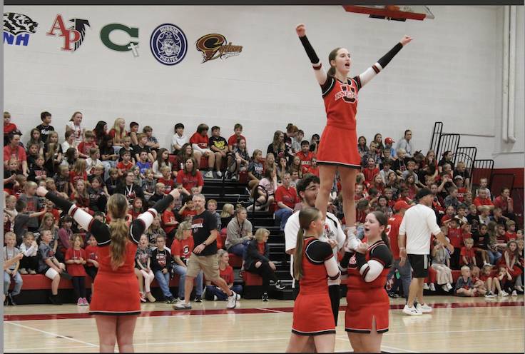 Cheerleaders in red and black uniforms perform a stunt during a school pep rally as students cheer from crowded gym bleachers.