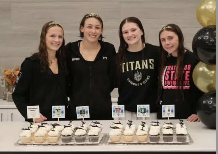 Four smiling student swimmers pose behind trays of decorated cupcakes at a celebration with black and gold balloons.