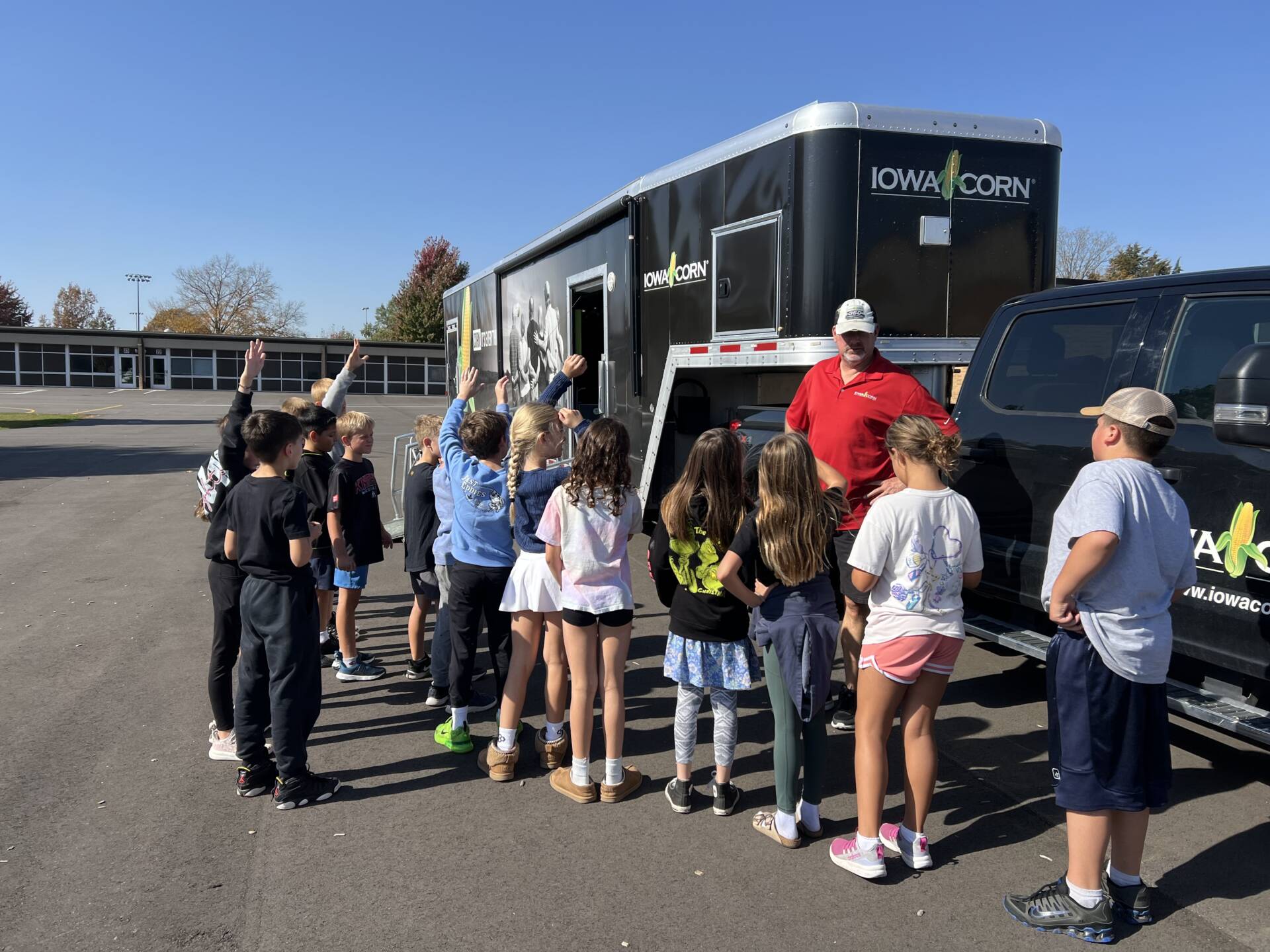 Students raise their hands while listening to a presenter beside an Iowa Corn educational trailer parked outside their school.
