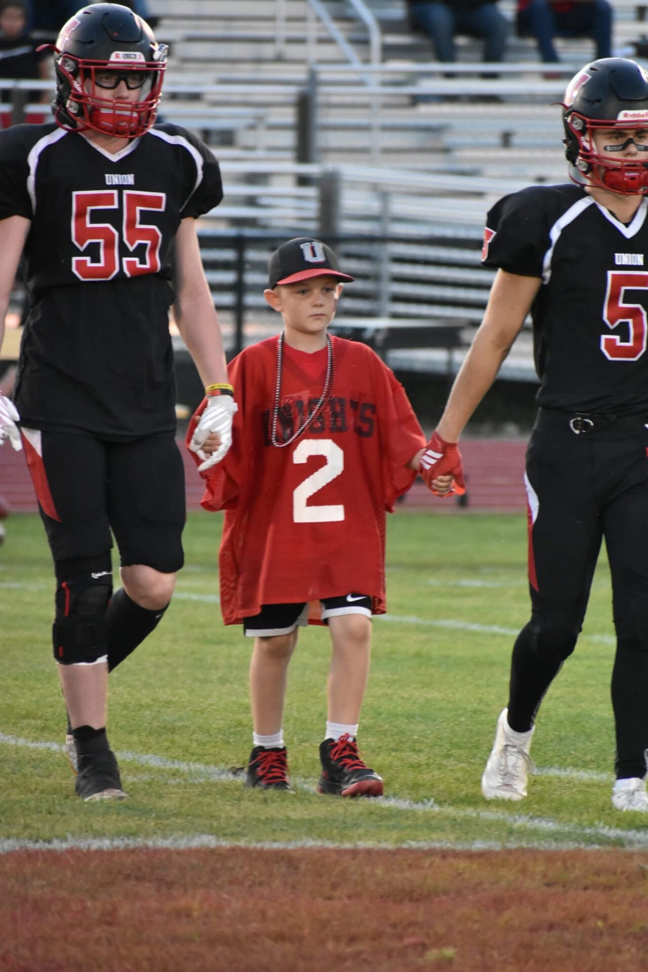 Young child in oversized red jersey walking hand in hand with football players.