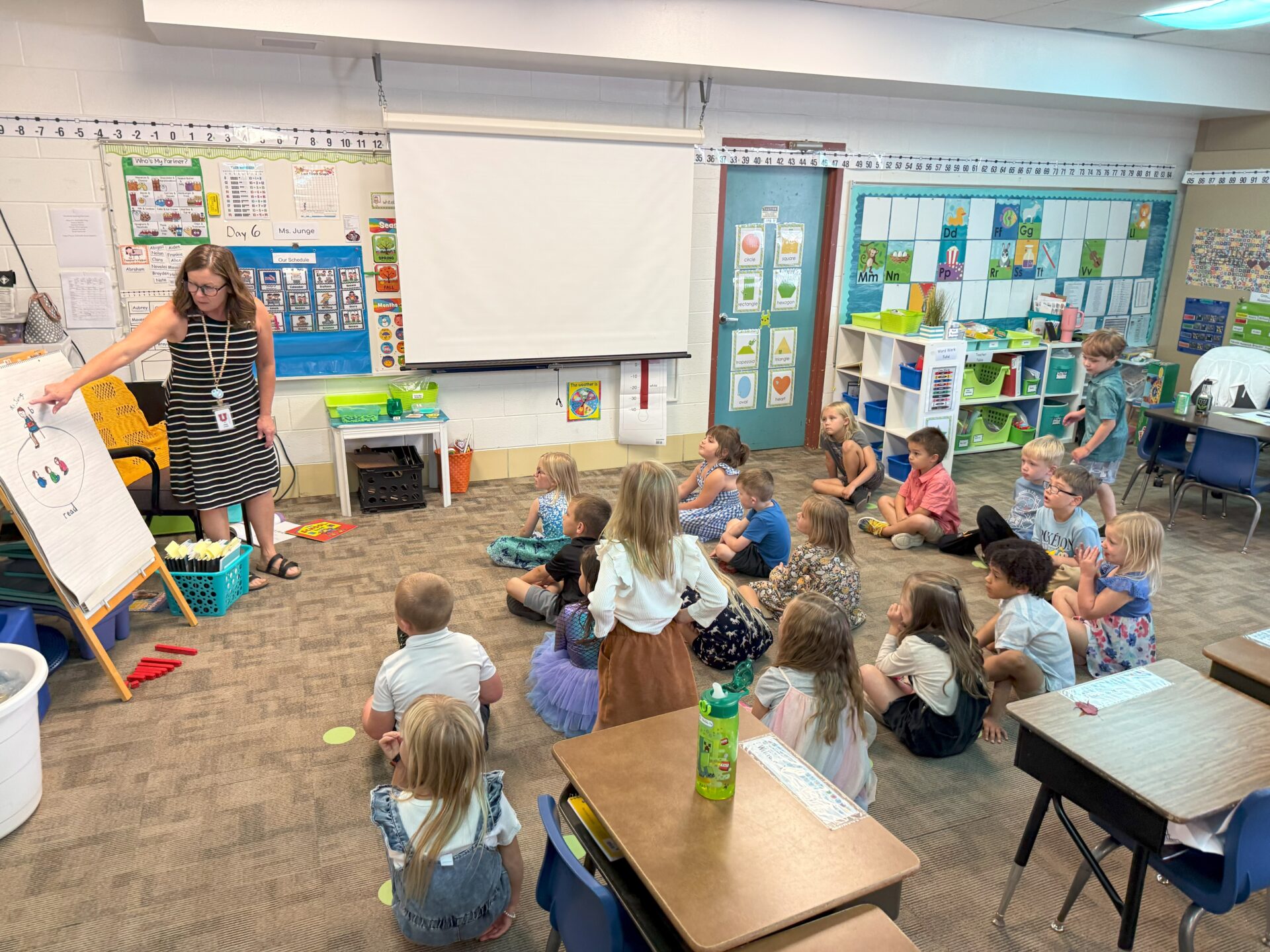 Teacher points to a chart at the front of the classroom while young students sit on the floor watching attentively.