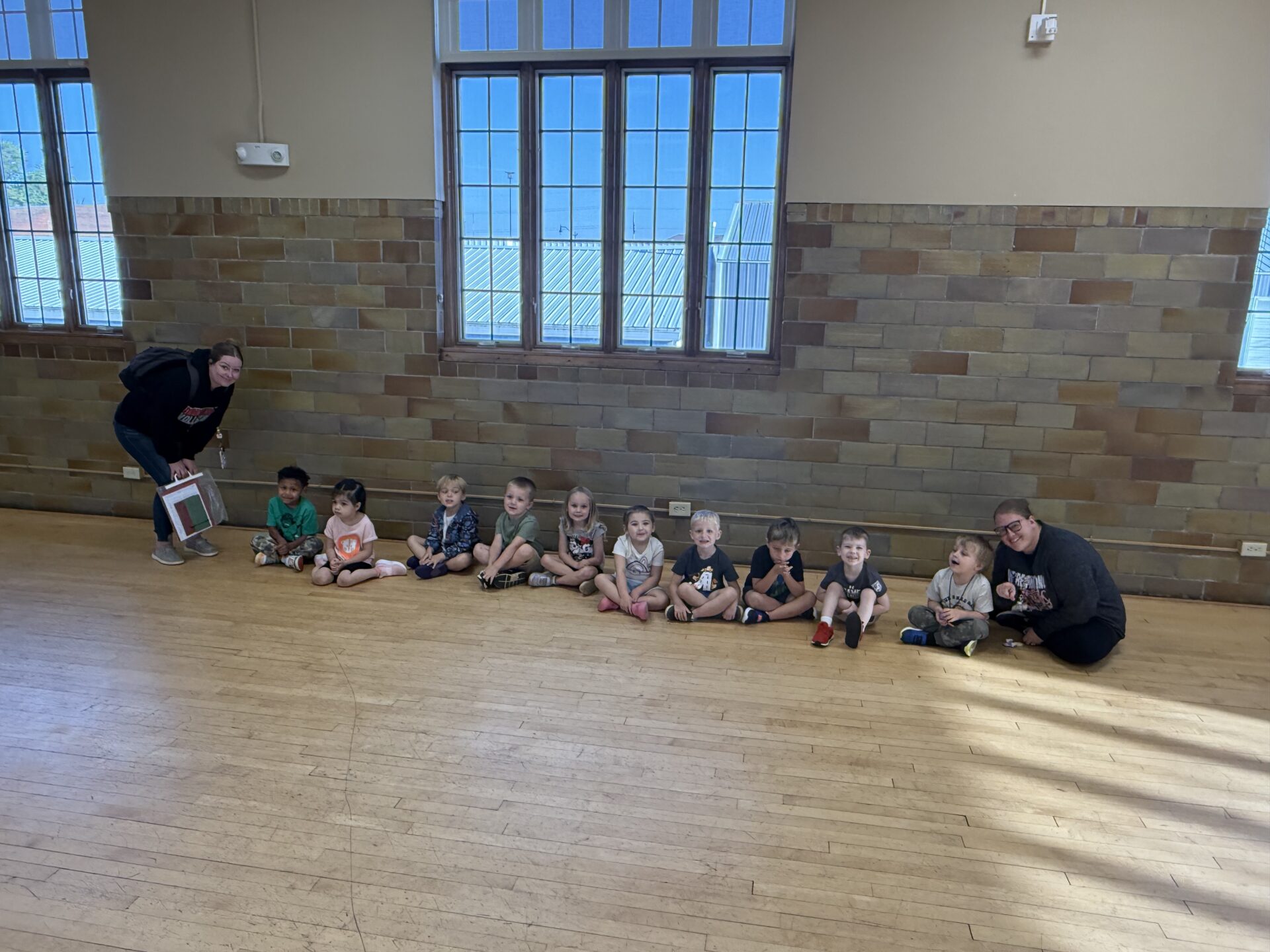 Two adults kneel at each end of a line of young children sitting on a gym floor in front of tall windows. The children smile and look toward the camera.