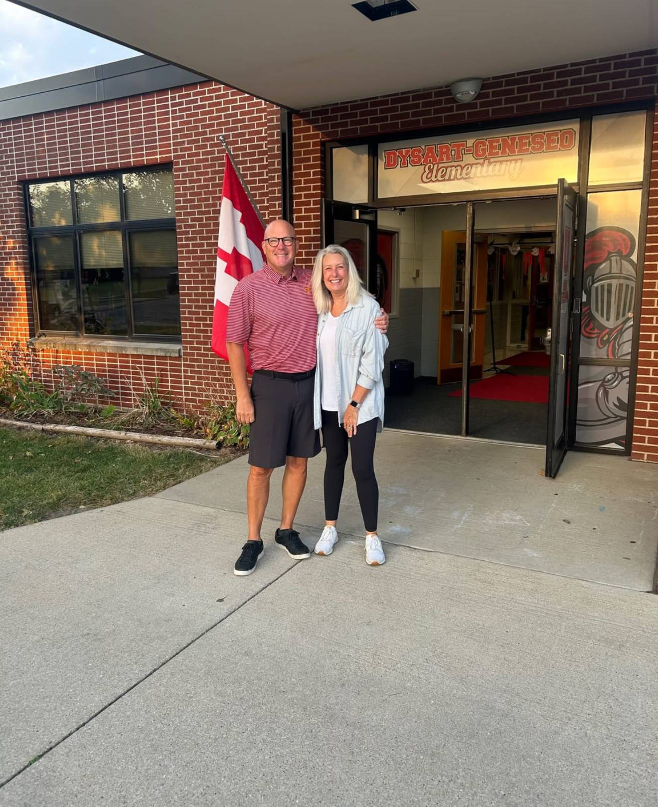 Two adults smile while standing together outside the entrance of Dysart-Geneseo Elementary.