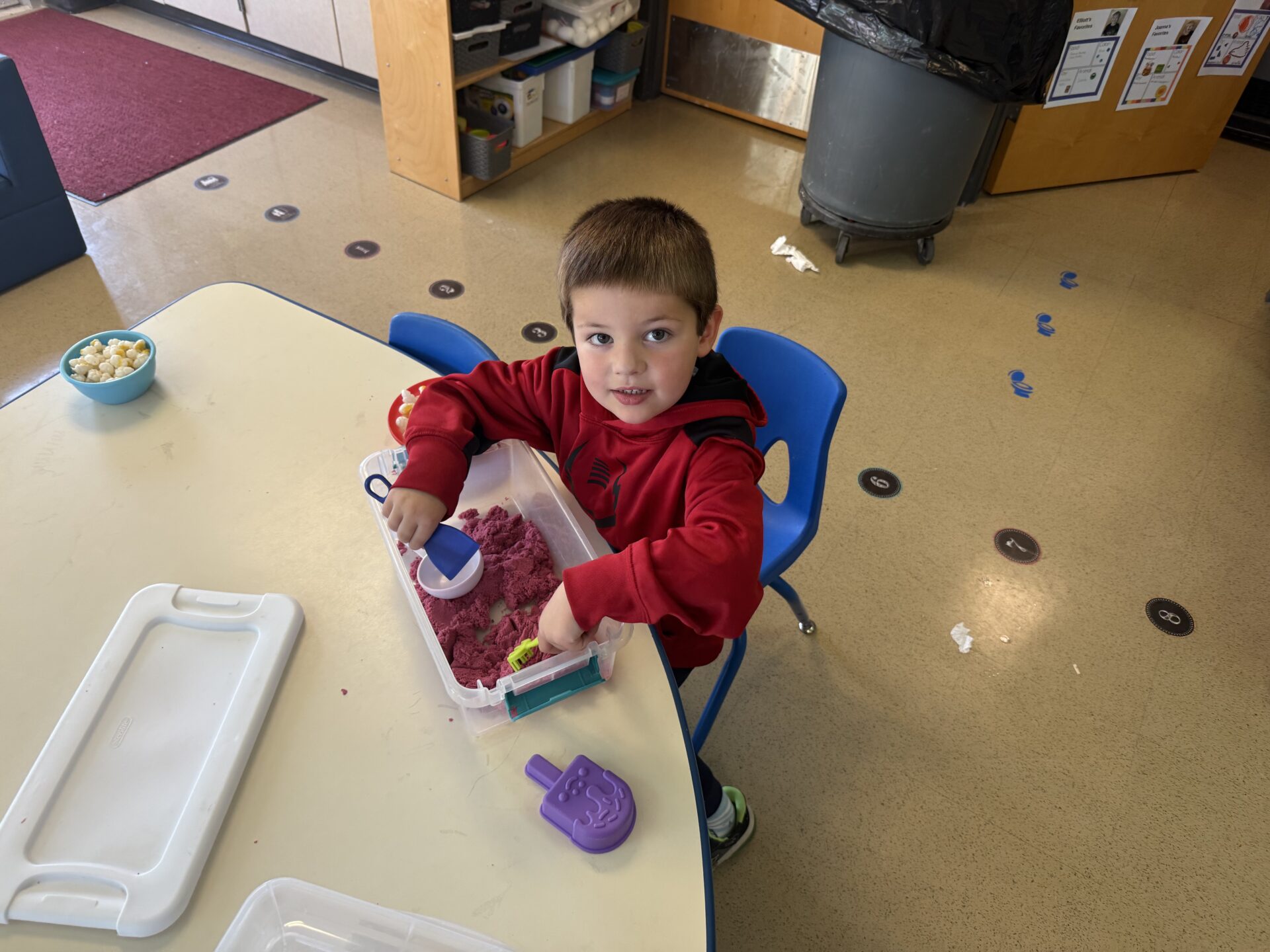 A preschool-aged student wearing a red hoodie plays with purple kinetic sand at a classroom table.