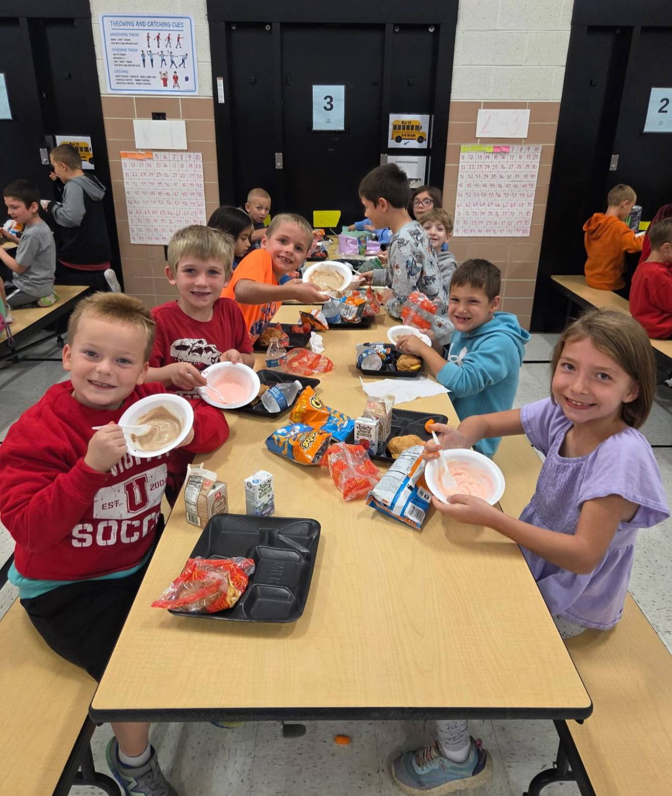 A group of elementary students smile at a cafeteria table while eating lunch. They hold bowls of pudding and snacks, with cartons of milk and juice on their trays.