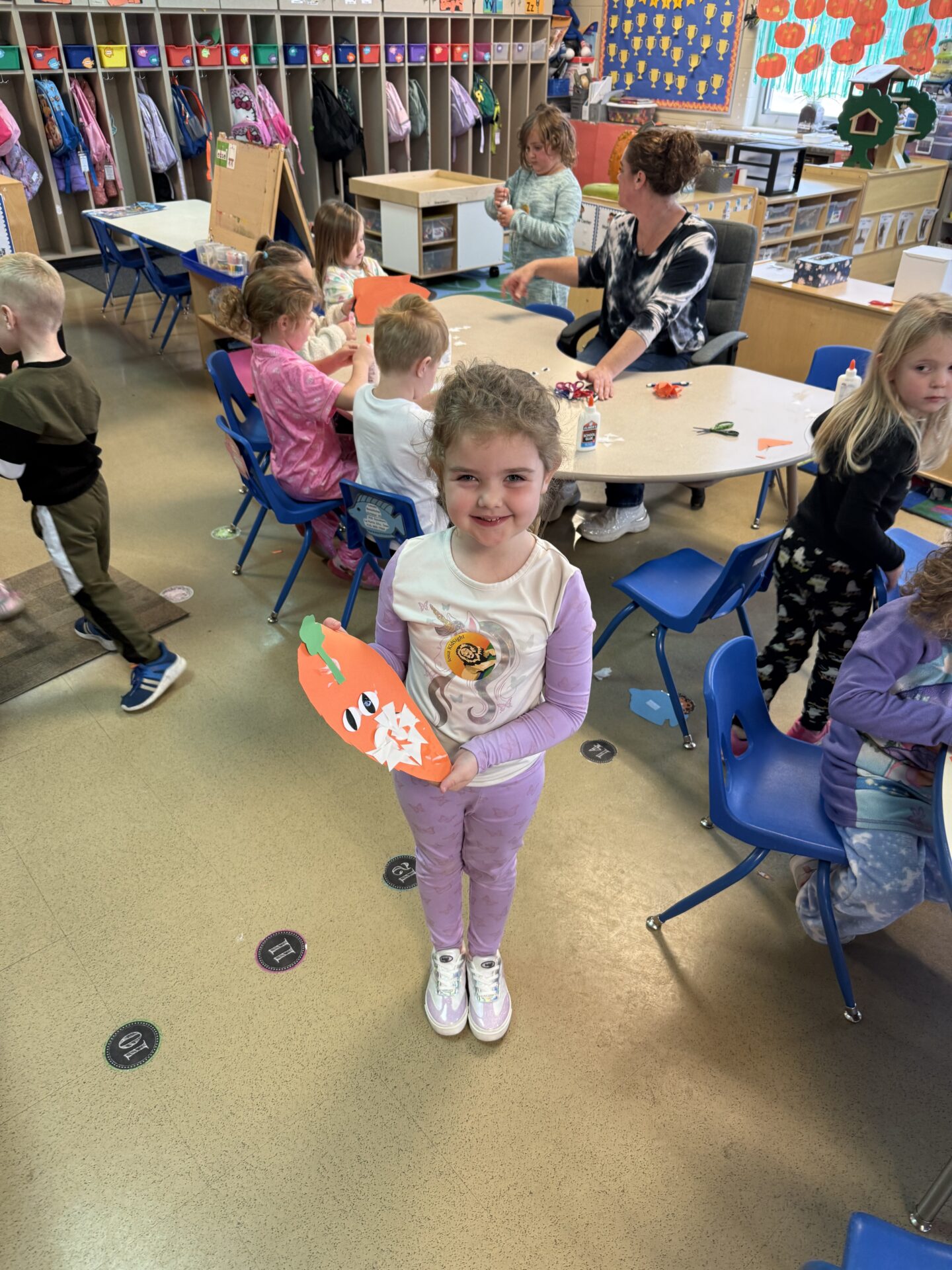 A young student in a light purple outfit smiles while holding a pumpkin-themed paper craft in a classroom.