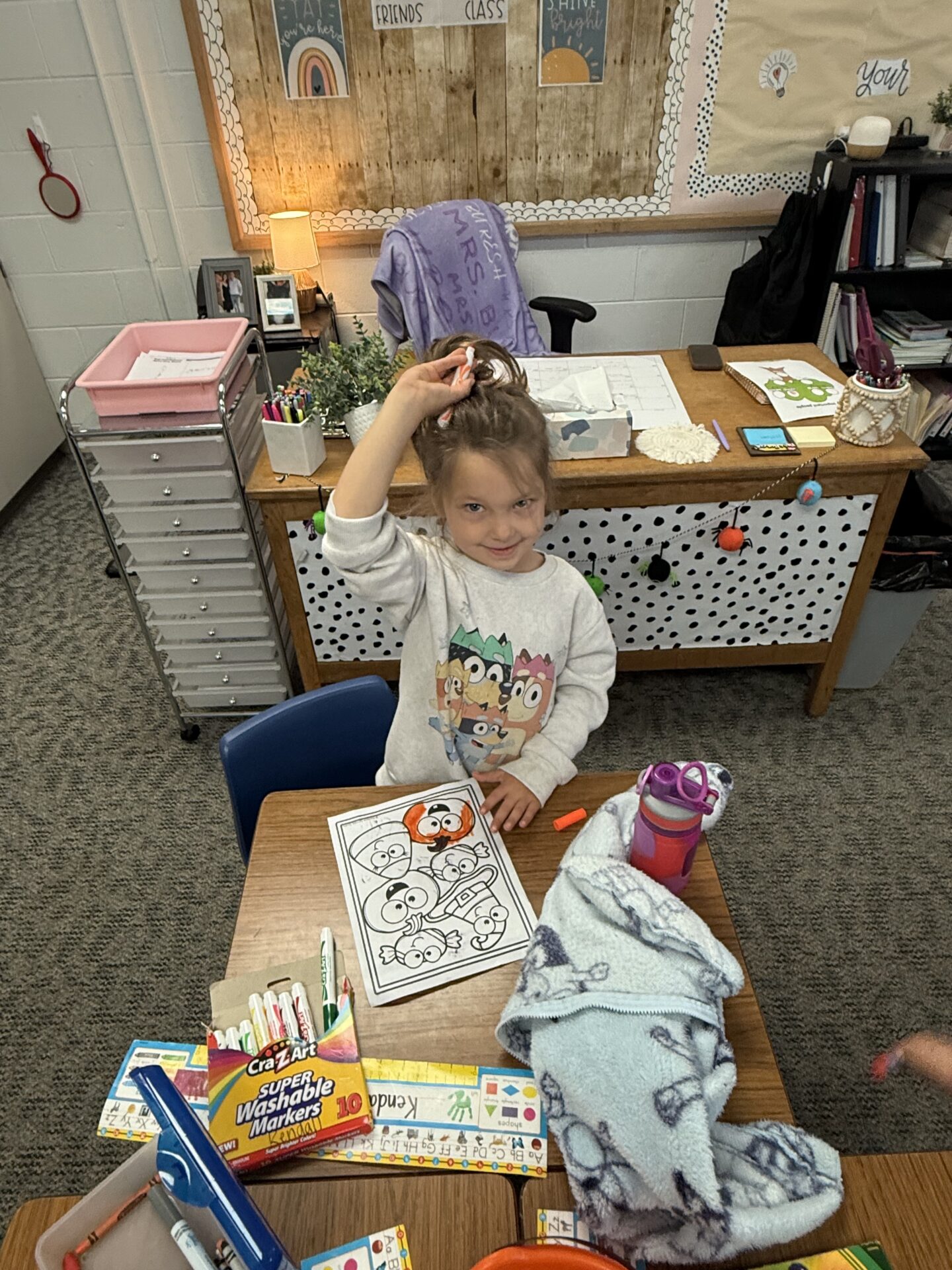 A smiling young student holds a marker and shows off her coloring sheet at her desk.