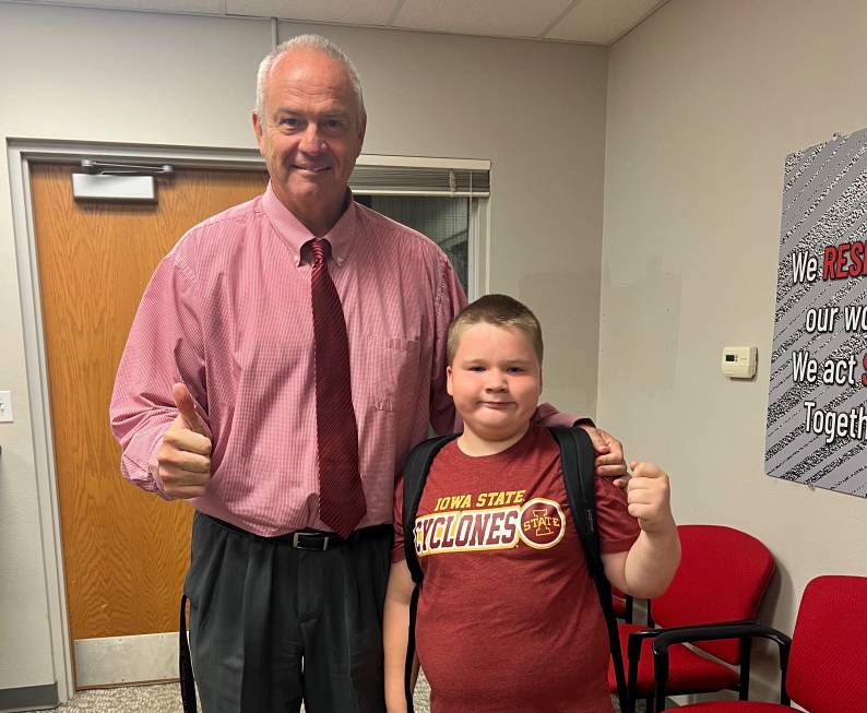 An adult and a student, both wearing red shirts, stand side by side giving thumbs up. The student’s shirt reads “Iowa State Cyclones,” and the two pose inside a school office area with red chairs.