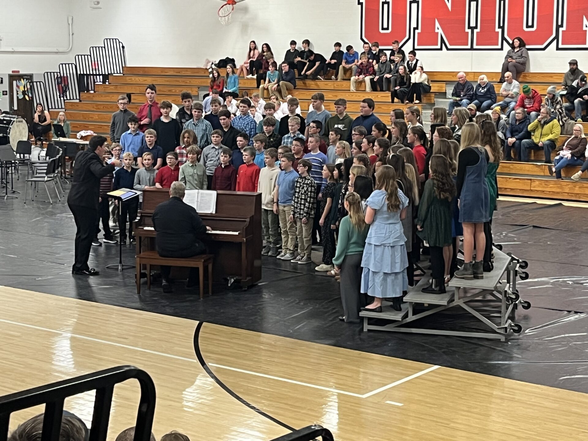 A large group of middle school students performs a choir concert in a gymnasium. A conductor leads them while a pianist plays at an upright piano.