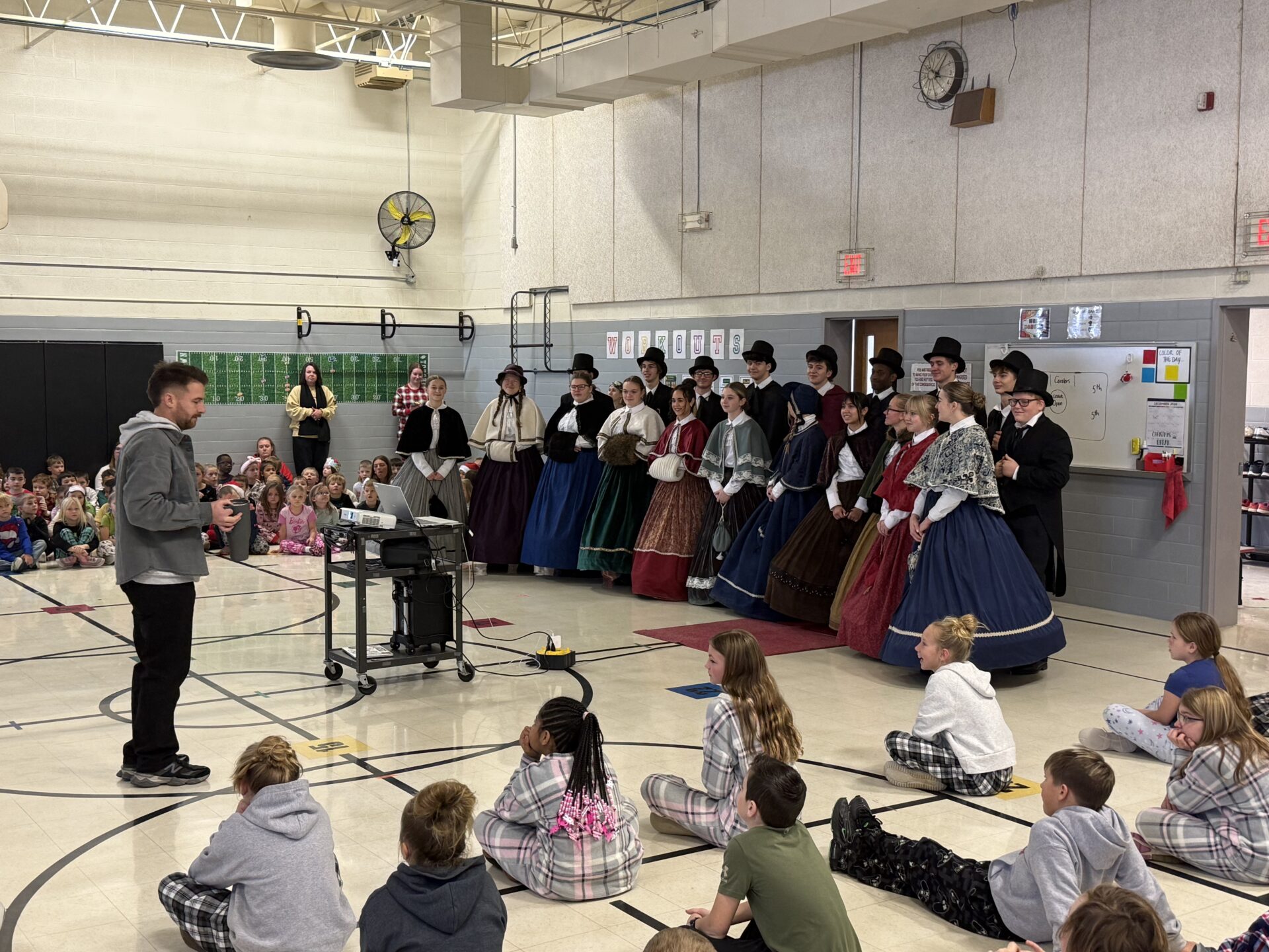 A group of high school students in Victorian costumes visits an elementary school gym. The students stand in front of seated elementary children, who are wearing pajamas.