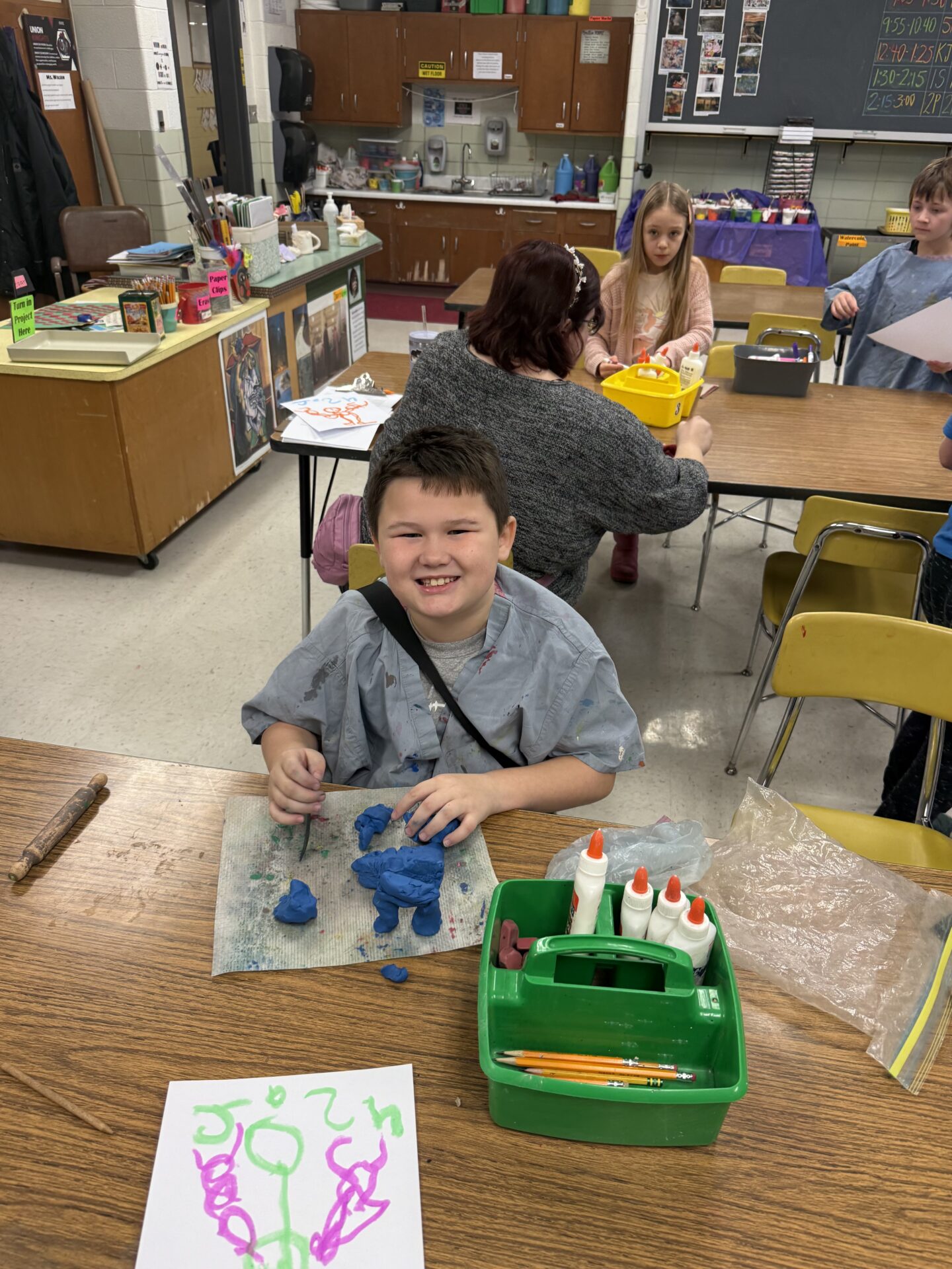 Smiling student works with blue modeling clay at a classroom table, surrounded by art supplies and a colorful drawing.