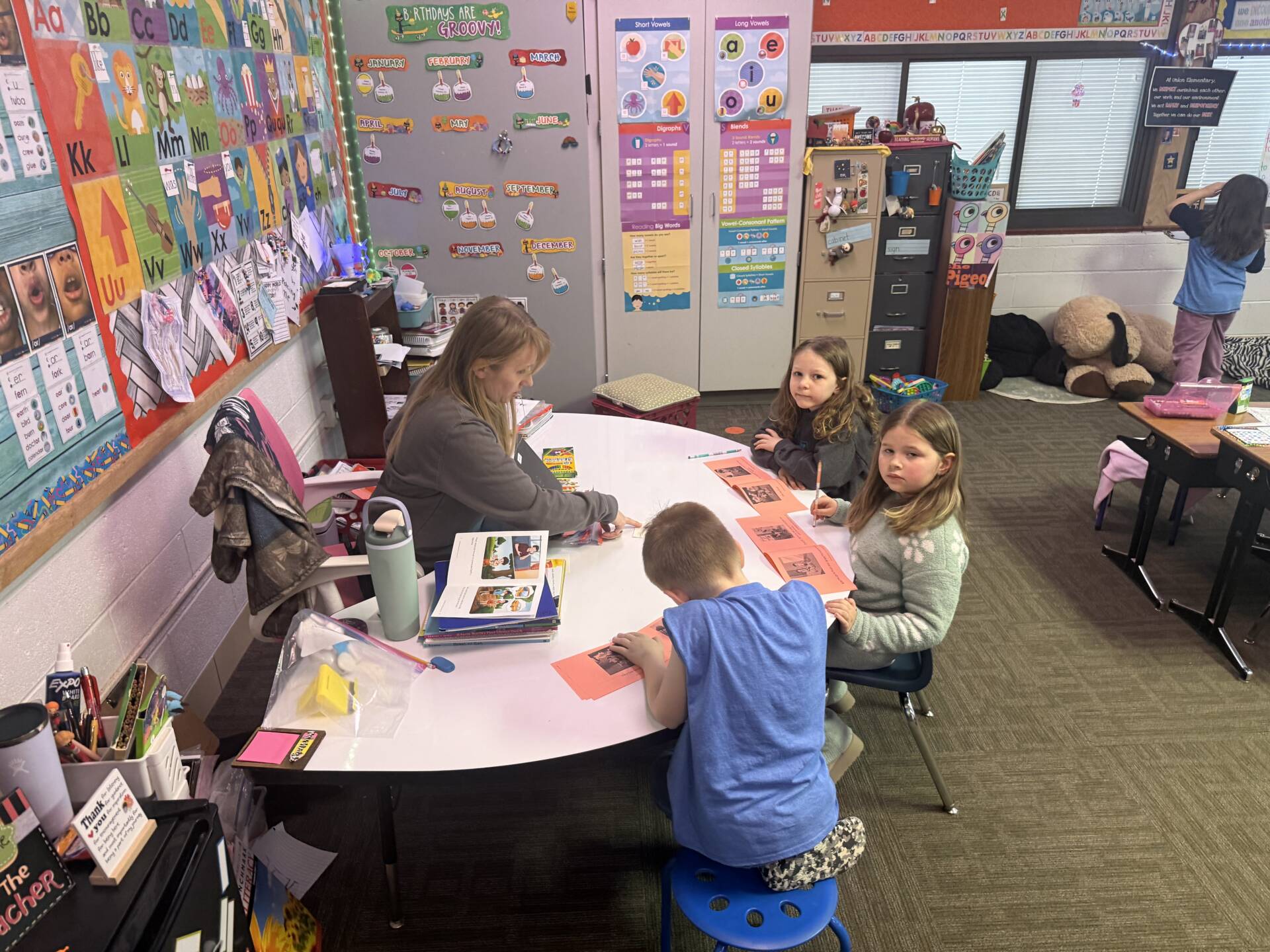A teacher works with three young students at a small table during a classroom activity while another student works nearby.
