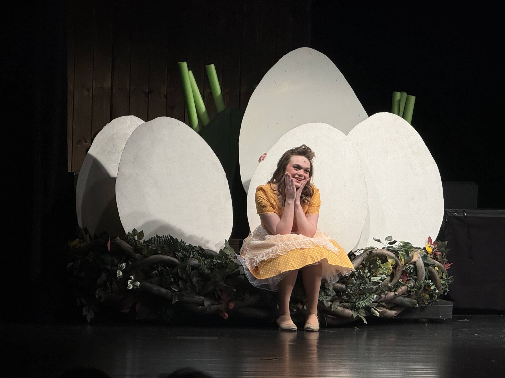 A student actor sits smiling among large egg props as part of a theatrical set during a school play.
