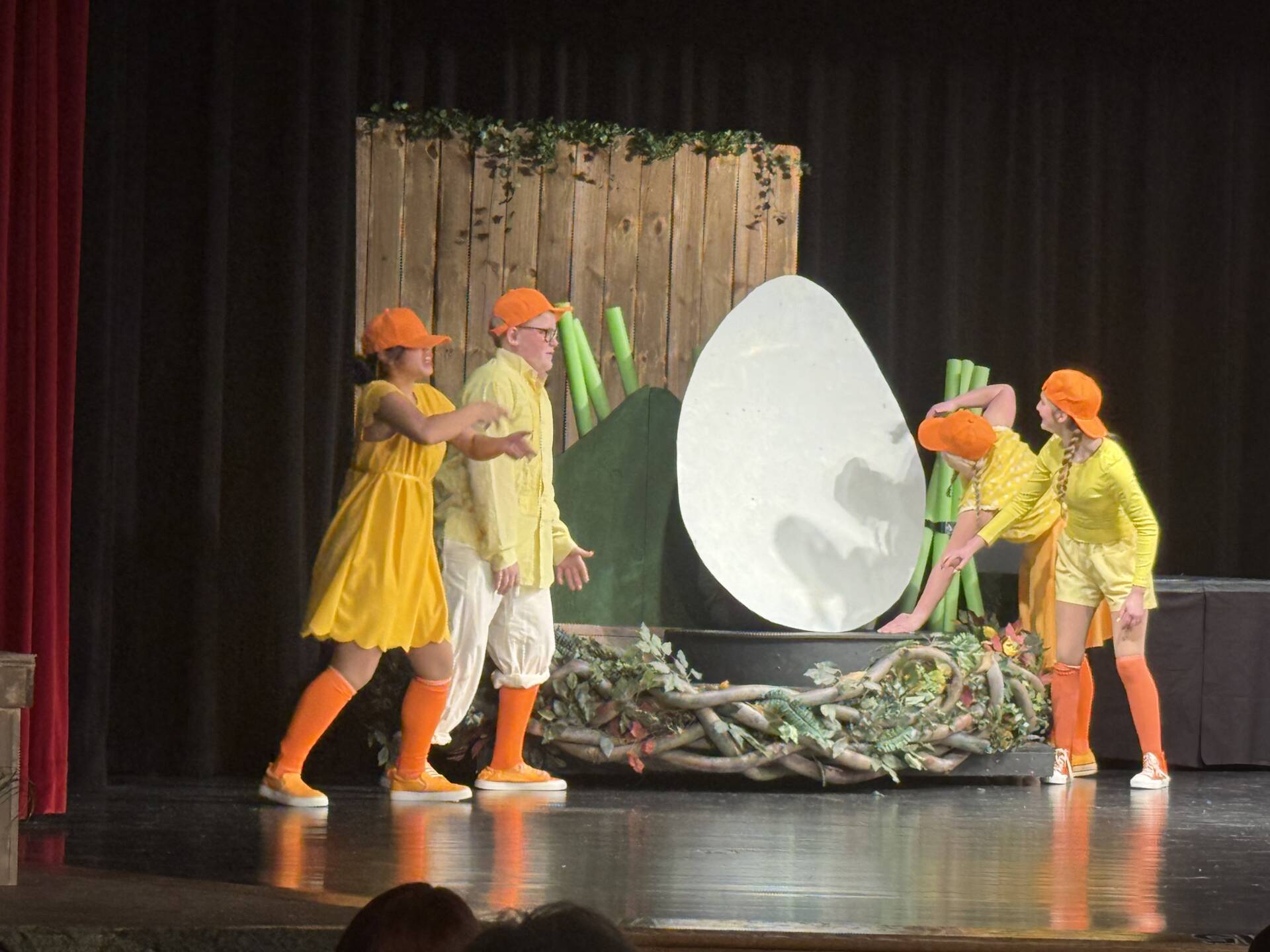 Student performers dressed in yellow costumes and orange hats act out a scene around a large prop egg during a stage production.