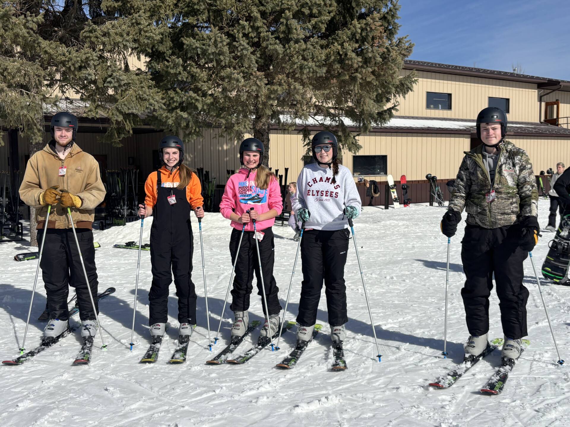 Five students wearing helmets stand on skis in the snow outside a lodge, holding ski poles during a school ski trip.