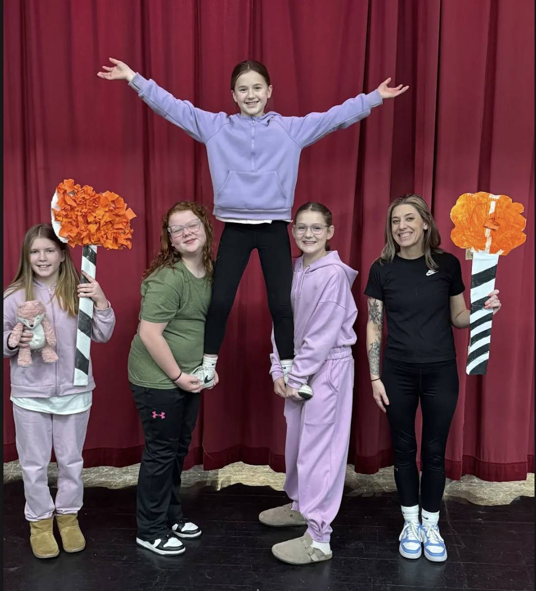 Four students and a teacher pose on a stage in front of red curtains while one student is lifted in a cheer pose and others hold orange pom-pom props.