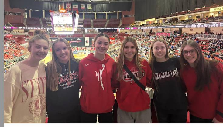 Six students smile for a photo at a basketball arena while attending a sporting event, with the court and crowd visible behind them.