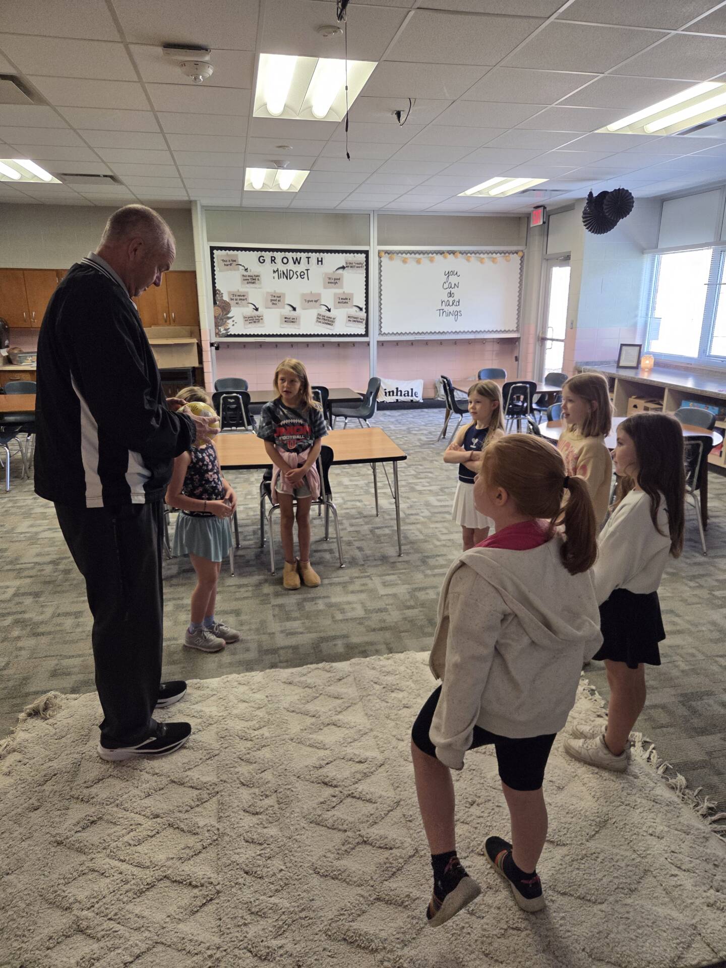 Teacher stands with a small group of students gathered on a rug, giving instructions during a classroom activity.