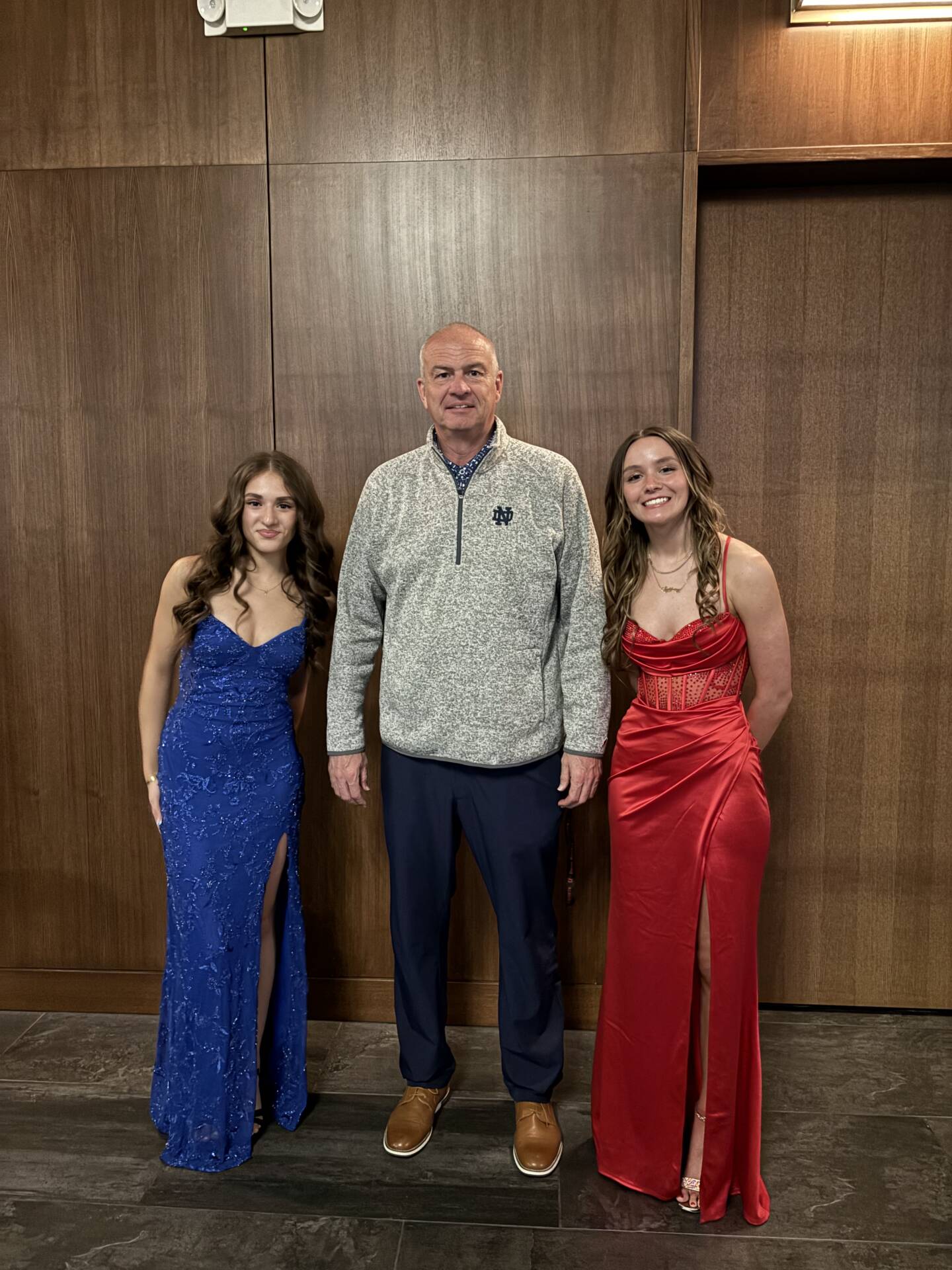 Two teens in formal dresses stand beside an adult man in casual attire, posing for a photo against a wood-paneled wall.
