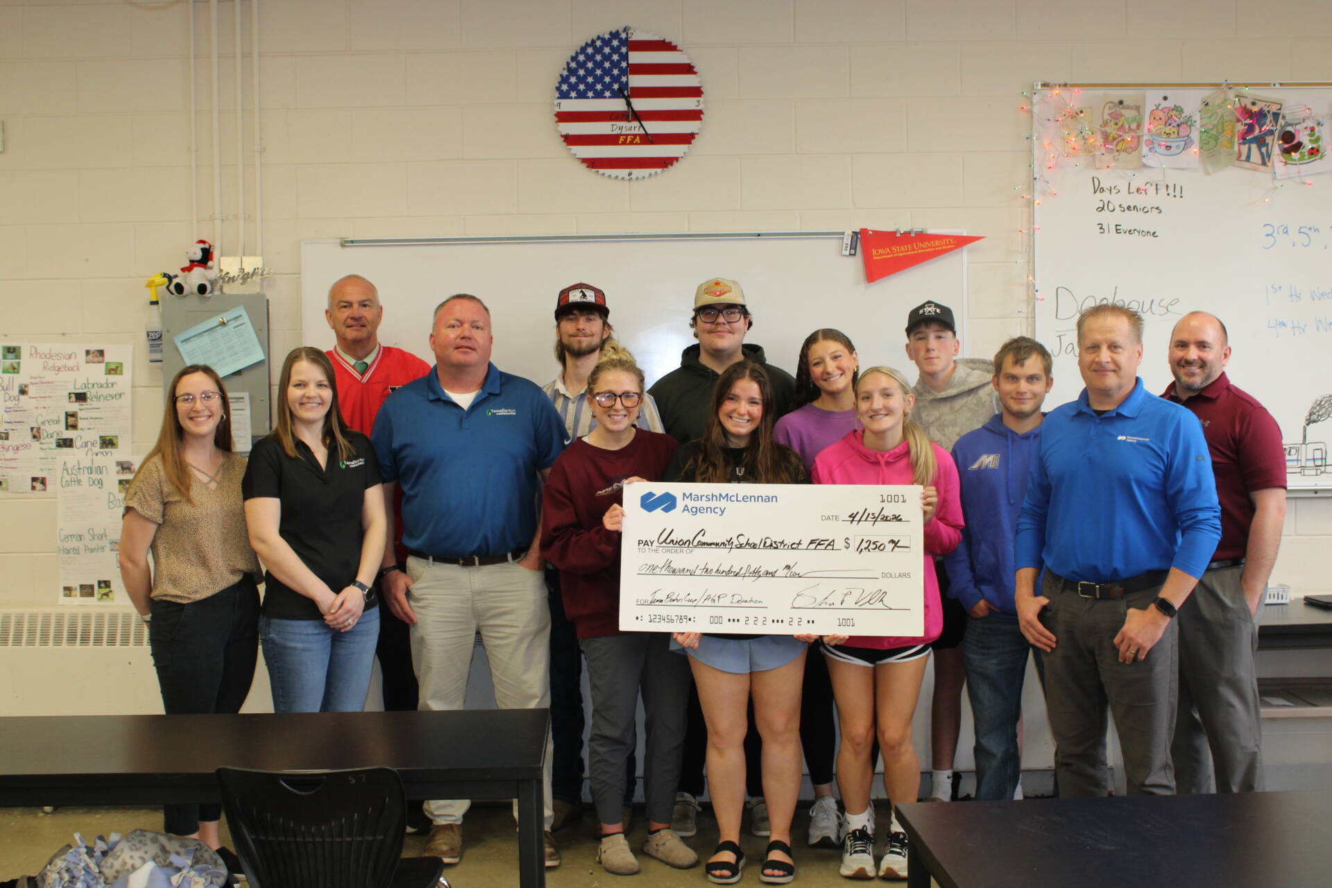 Group of students and adults stand in a classroom holding a large ceremonial check, smiling during a school recognition moment.