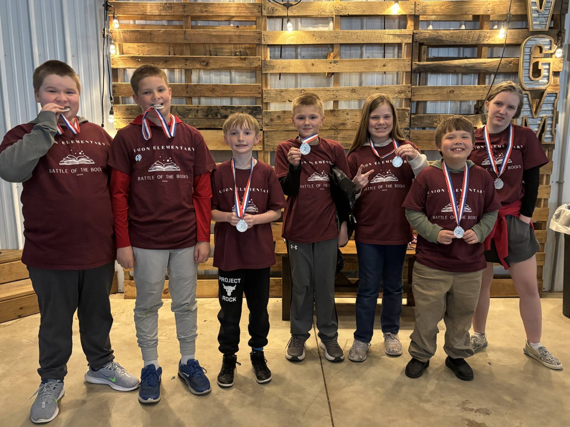 Seven students in maroon shirts stand together wearing medals, smiling and showing them to the camera.