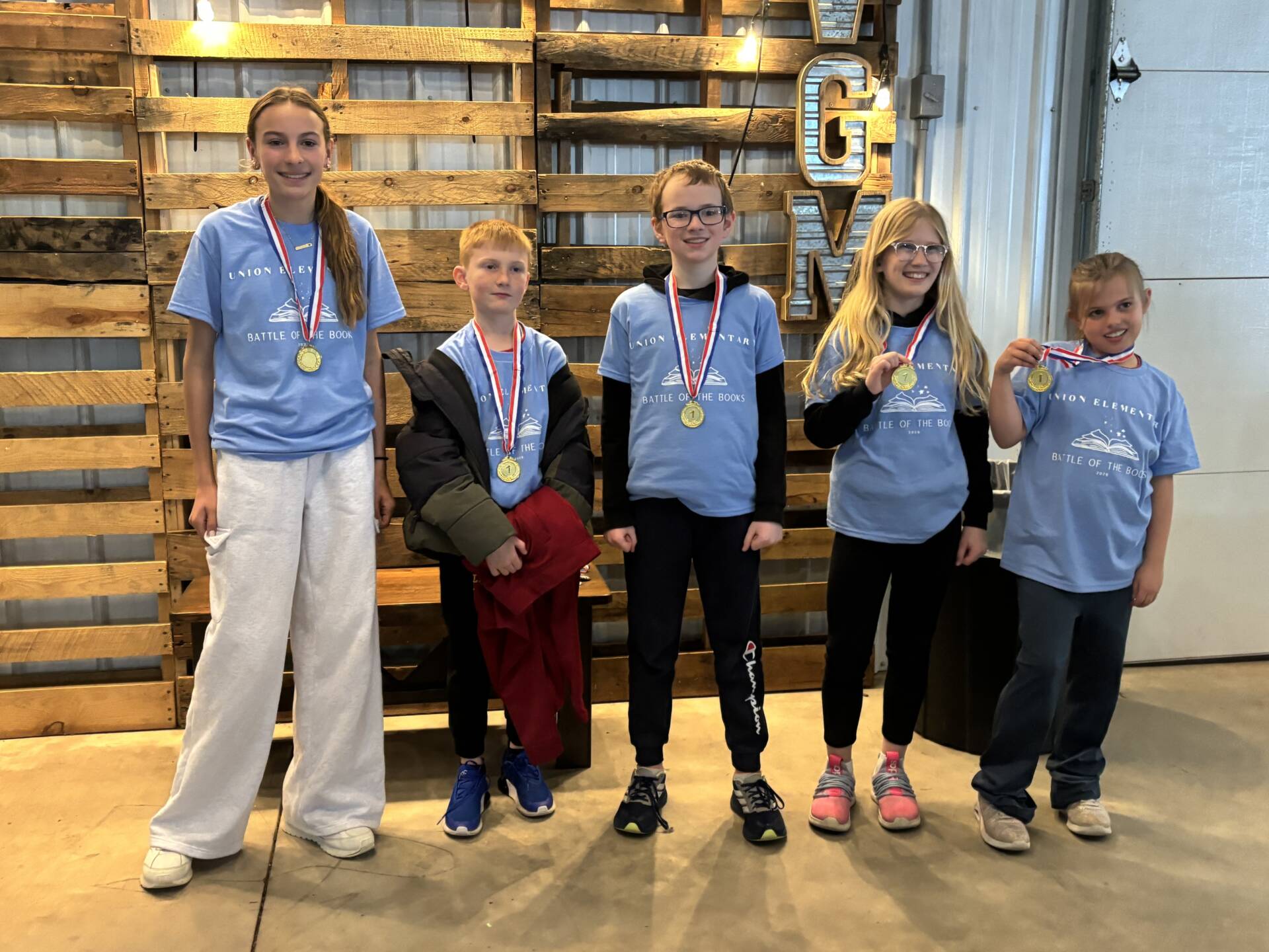 Five students in blue shirts stand with medals, smiling together in front of a wooden wall display.