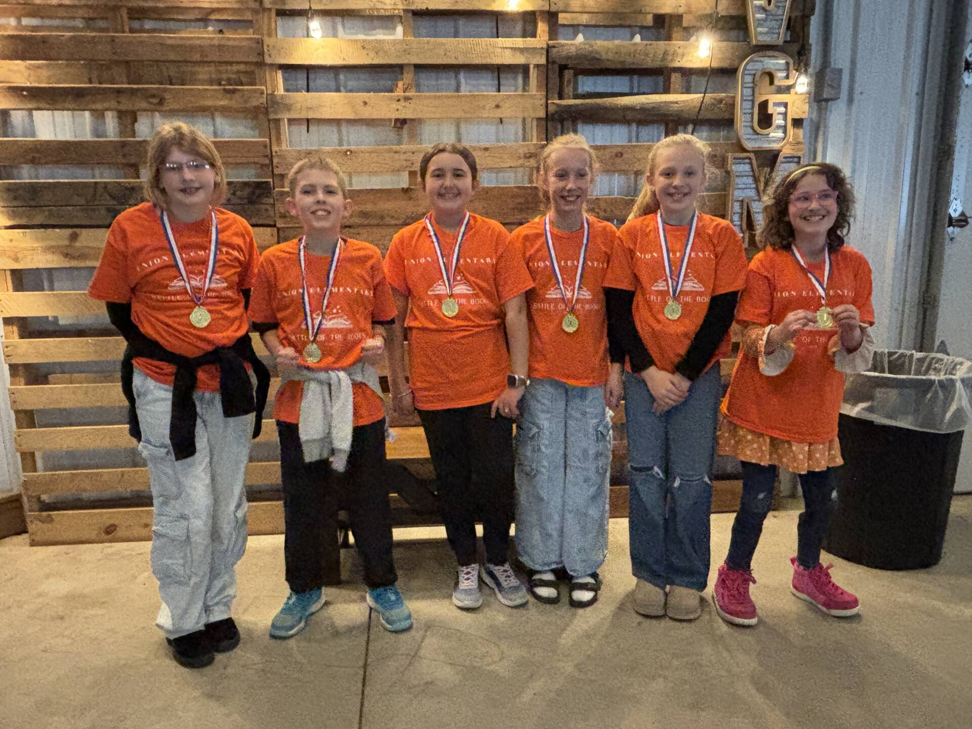 Six students in orange shirts stand in a row wearing medals, smiling in front of a wooden backdrop.