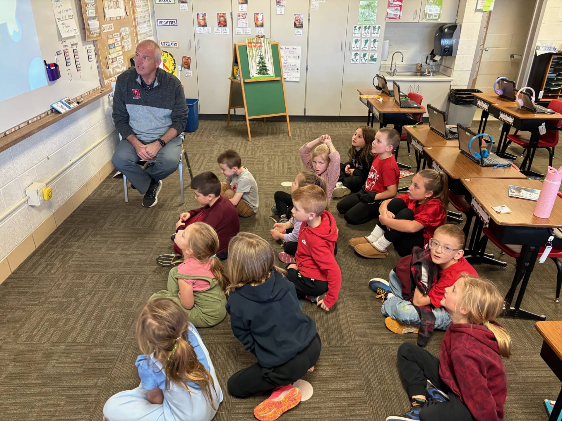 Adult sits at the front of a classroom speaking to students seated on the floor, who listen attentively.