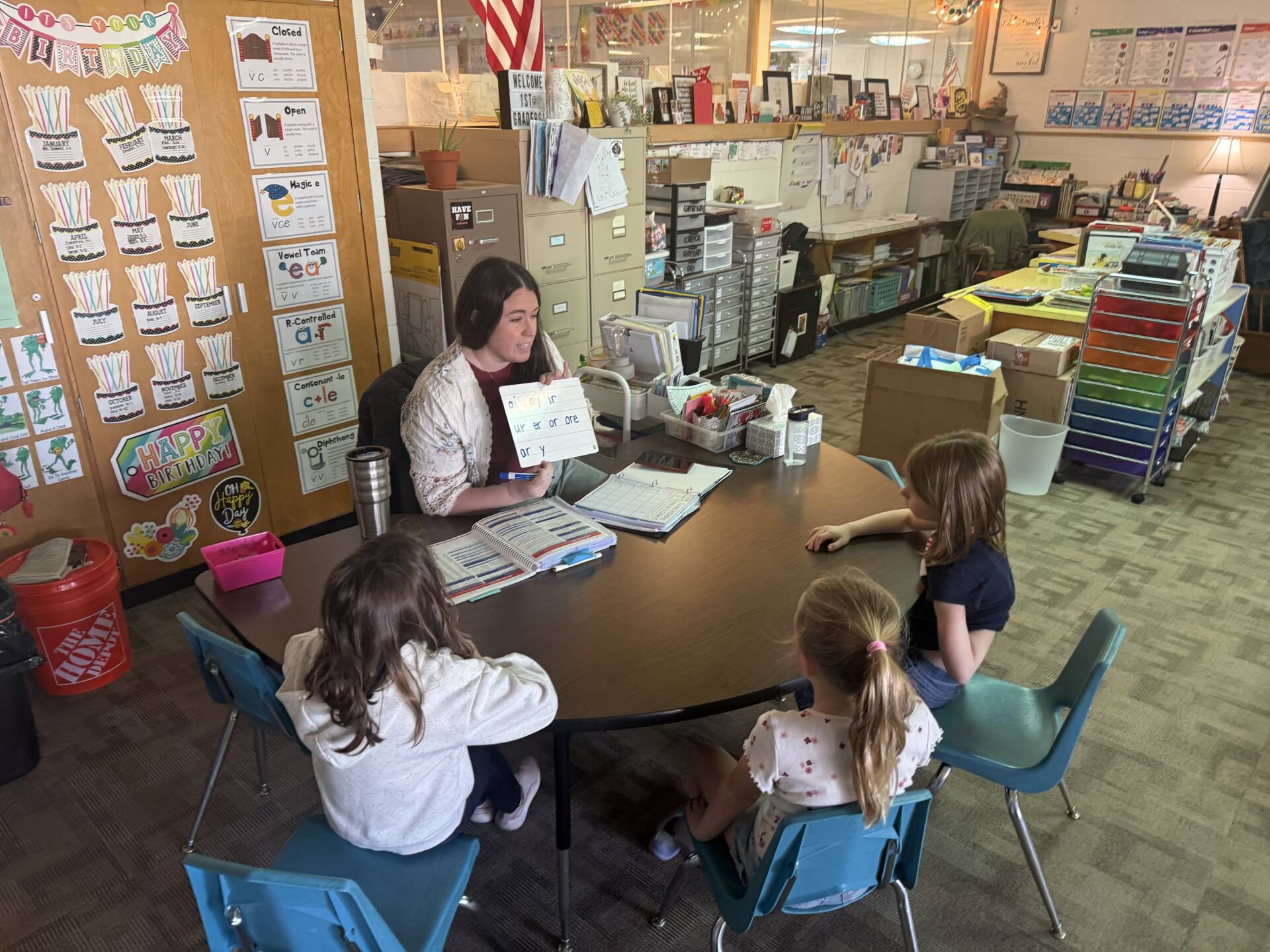 Teacher holds a phonics card while leading a small reading group of young students seated around a table.
