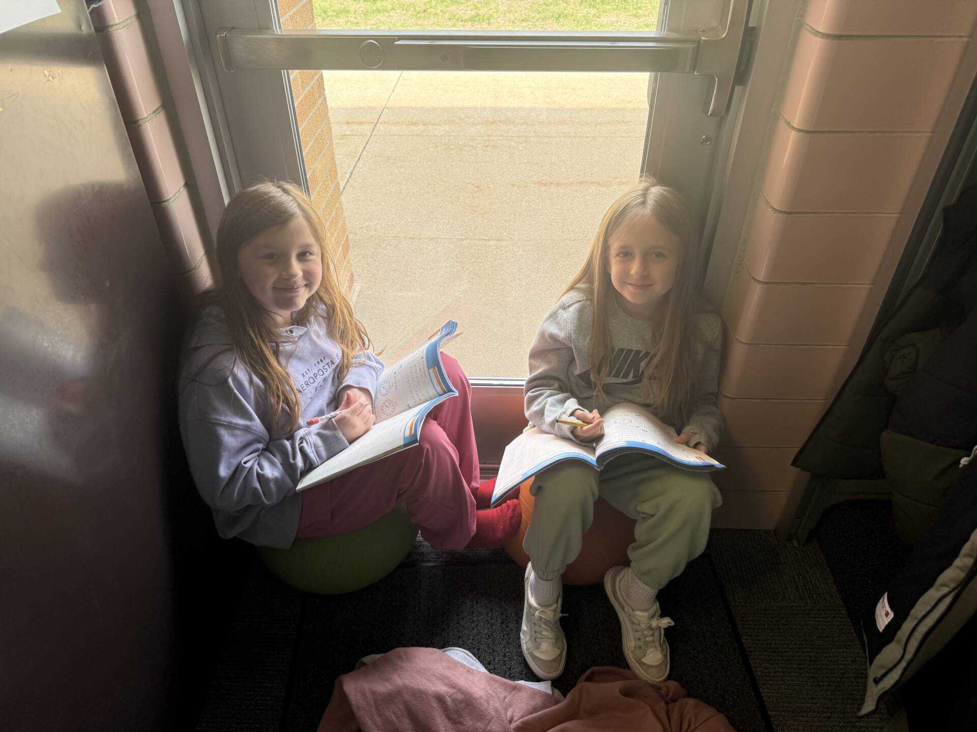 Two girls sit by a window on flexible seats, smiling while working in open notebooks.
