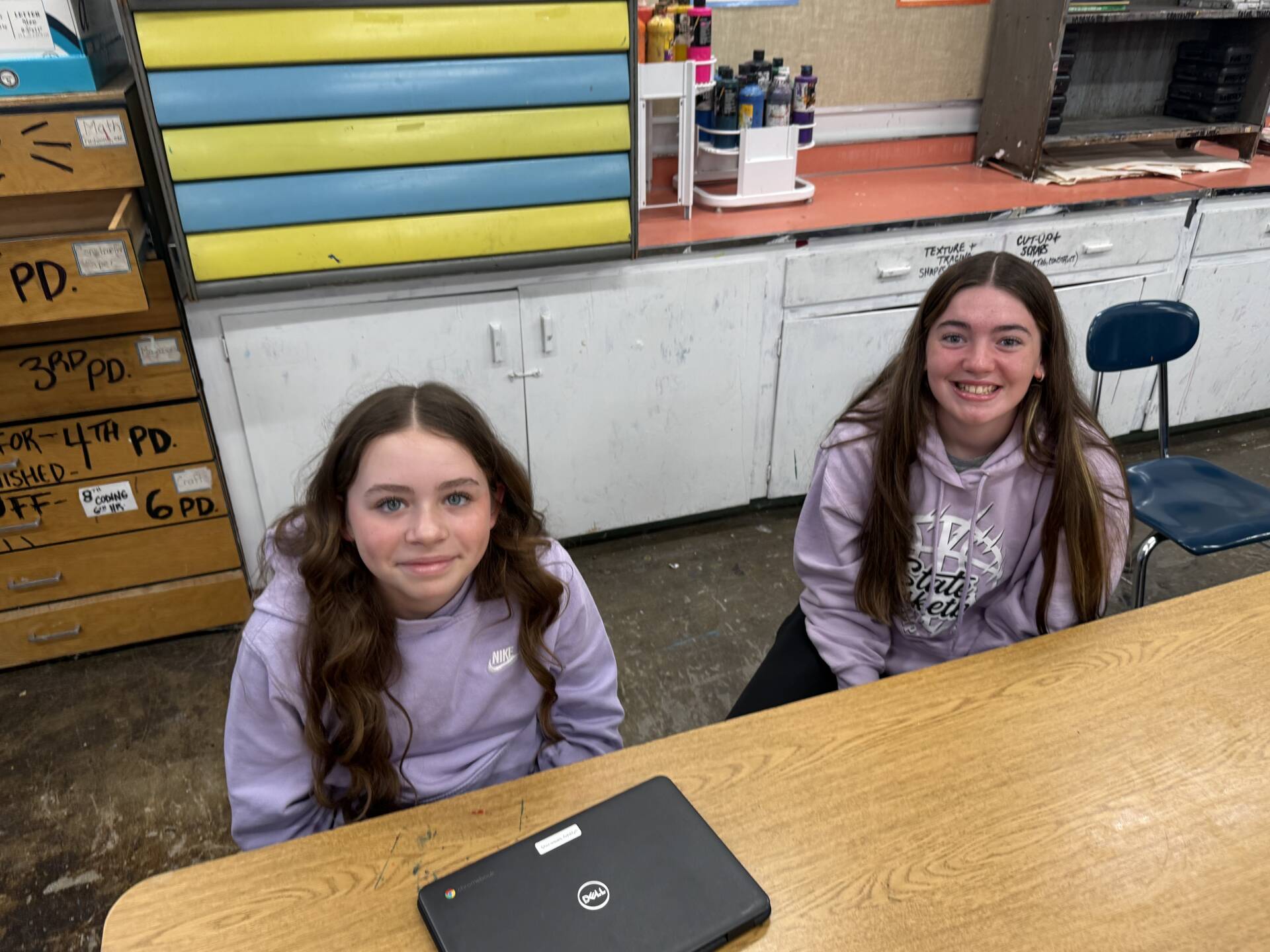 Two students sit at a classroom table, smiling near a laptop with art supplies and cabinets in the background.